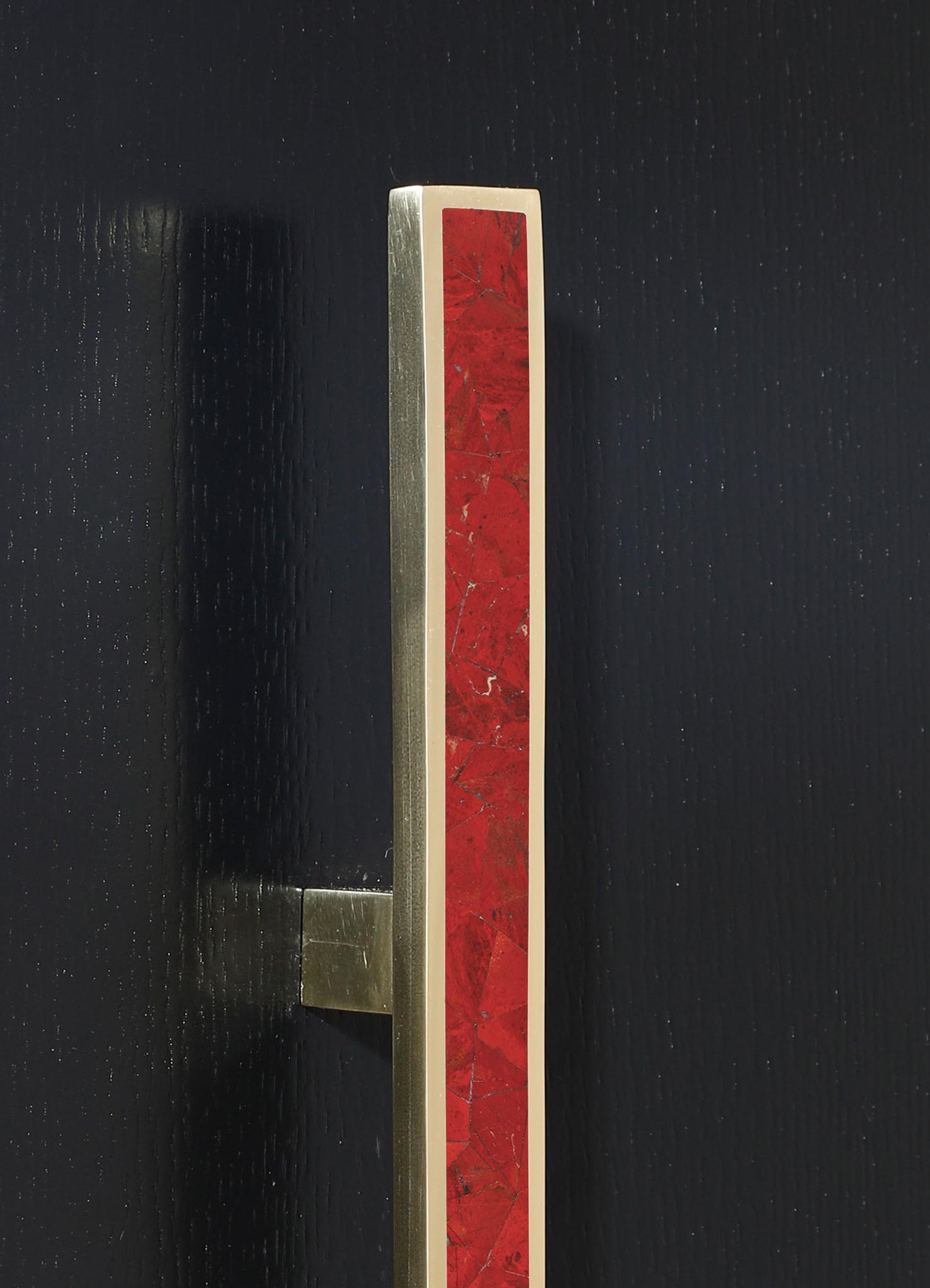 A vertical brass and wood door handle with a red mosaic tile insert, mounted on a black wooden door. The handle features clean, modern lines and a contemporary style.