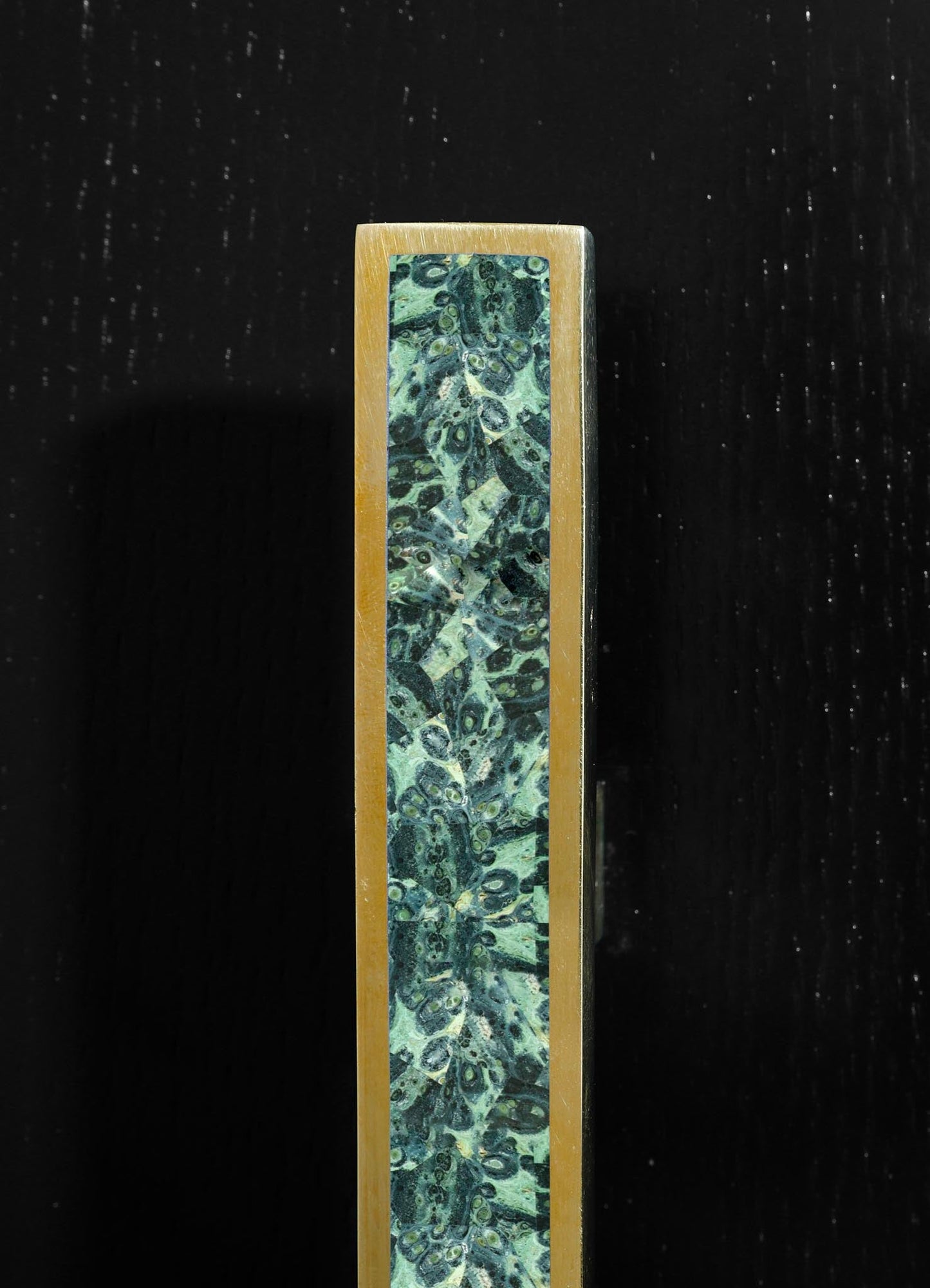 A rectangular object with a brass frame and a central panel featuring a green and black marbled pattern, possibly a decorative piece or small sculpture. The background is black with a textured surface.