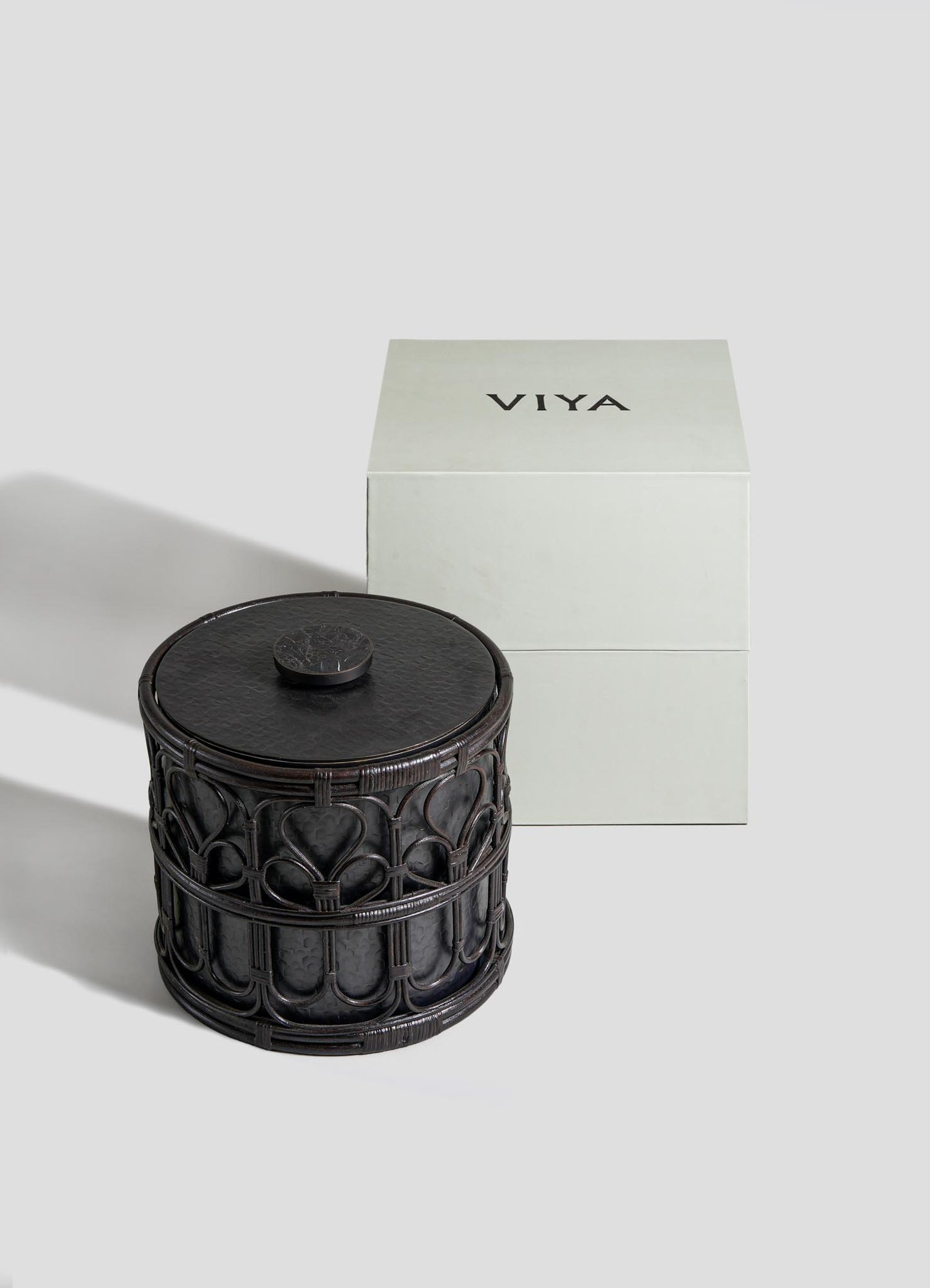 The VIYA Manipur Ice Bucket Large, a round, dark-toned container with a detailed hand-beaten brass design and lid, sits beside a simple white box labeled VIYA in bold black letters.