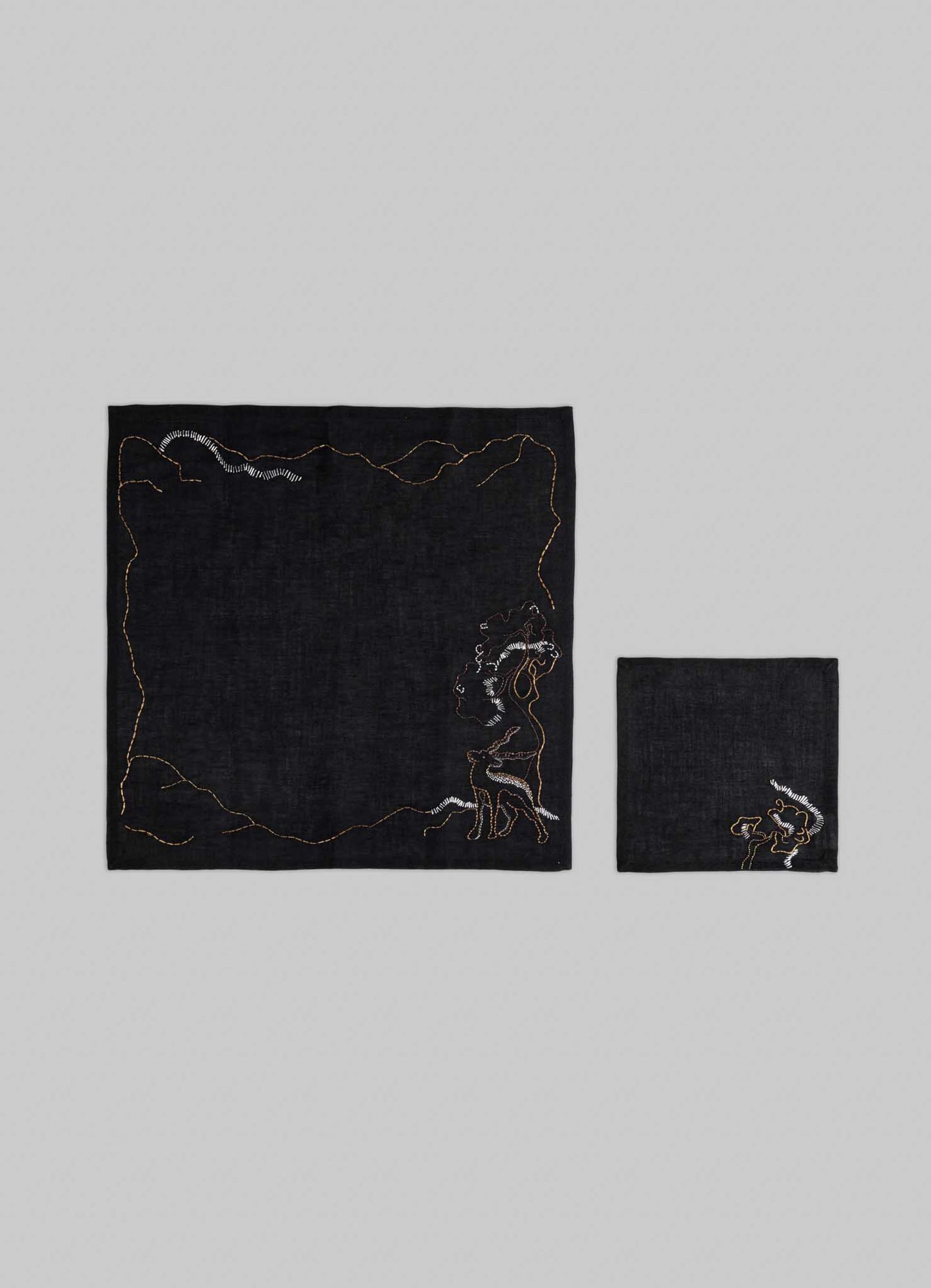 The Viya Blackbuck Dreams Hand Embroidered Black & Gold Cocktail Napkins (set of 4) feature unique manuscript-inspired motifs; shown are two napkins displayed on a gray background, one unfolded, the other folded.