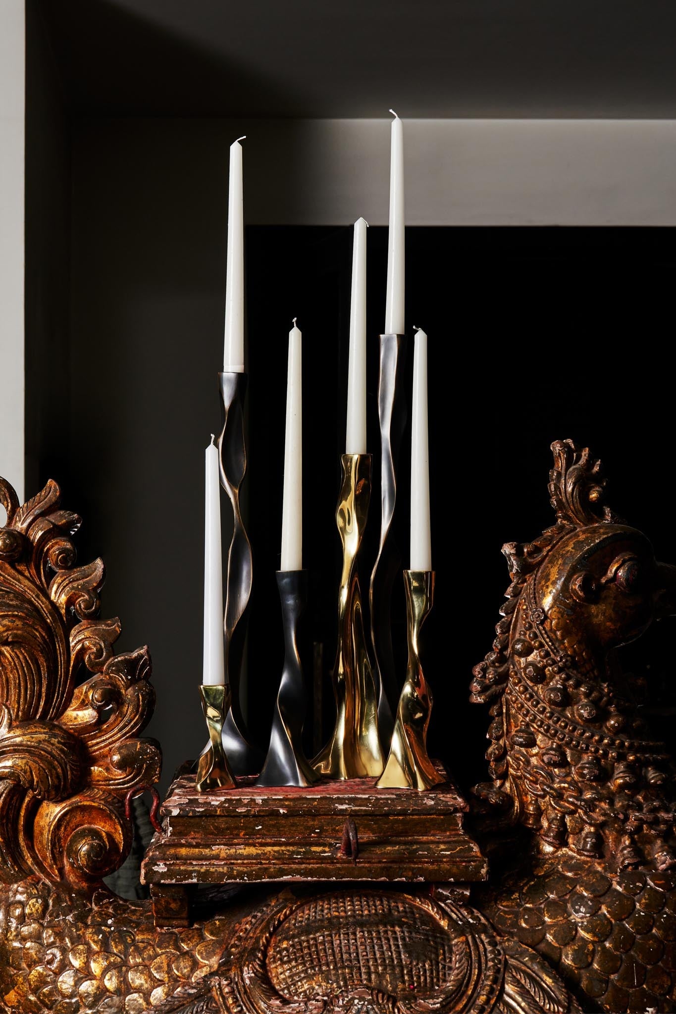 Tall taper candles with twisted metallic holders in black, gold, and silver finishes, displayed on a distressed wooden base, surrounded by ornate carved wooden decorative elements.