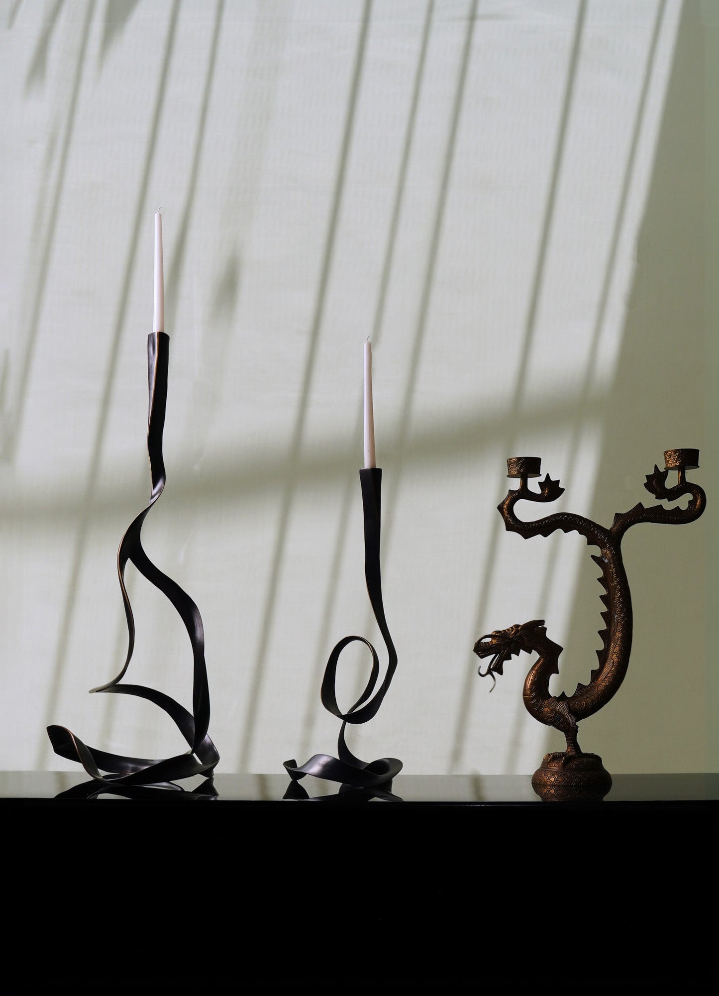 Decorative metal candle holders with twisted and sculptural designs, holding white tapered candles, placed on a dark surface against a light-colored wall with shadow patterns.