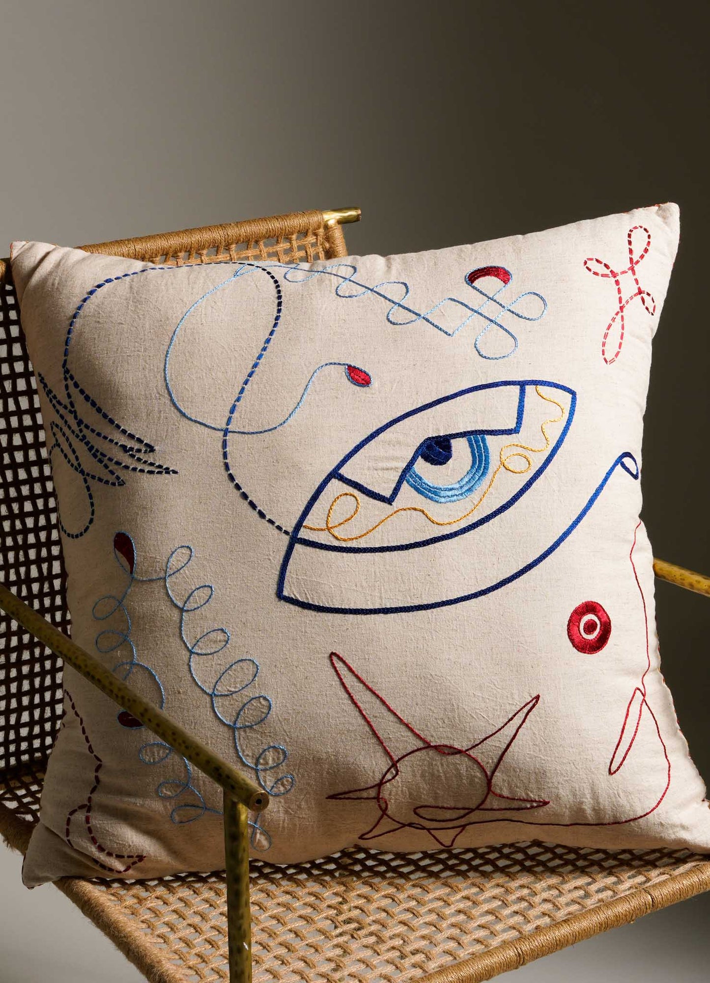Decorative square cushion with embroidered abstract facial motif in blue, red, and yellow thread on a cream fabric cover, placed on a woven rattan chair with a metal frame.