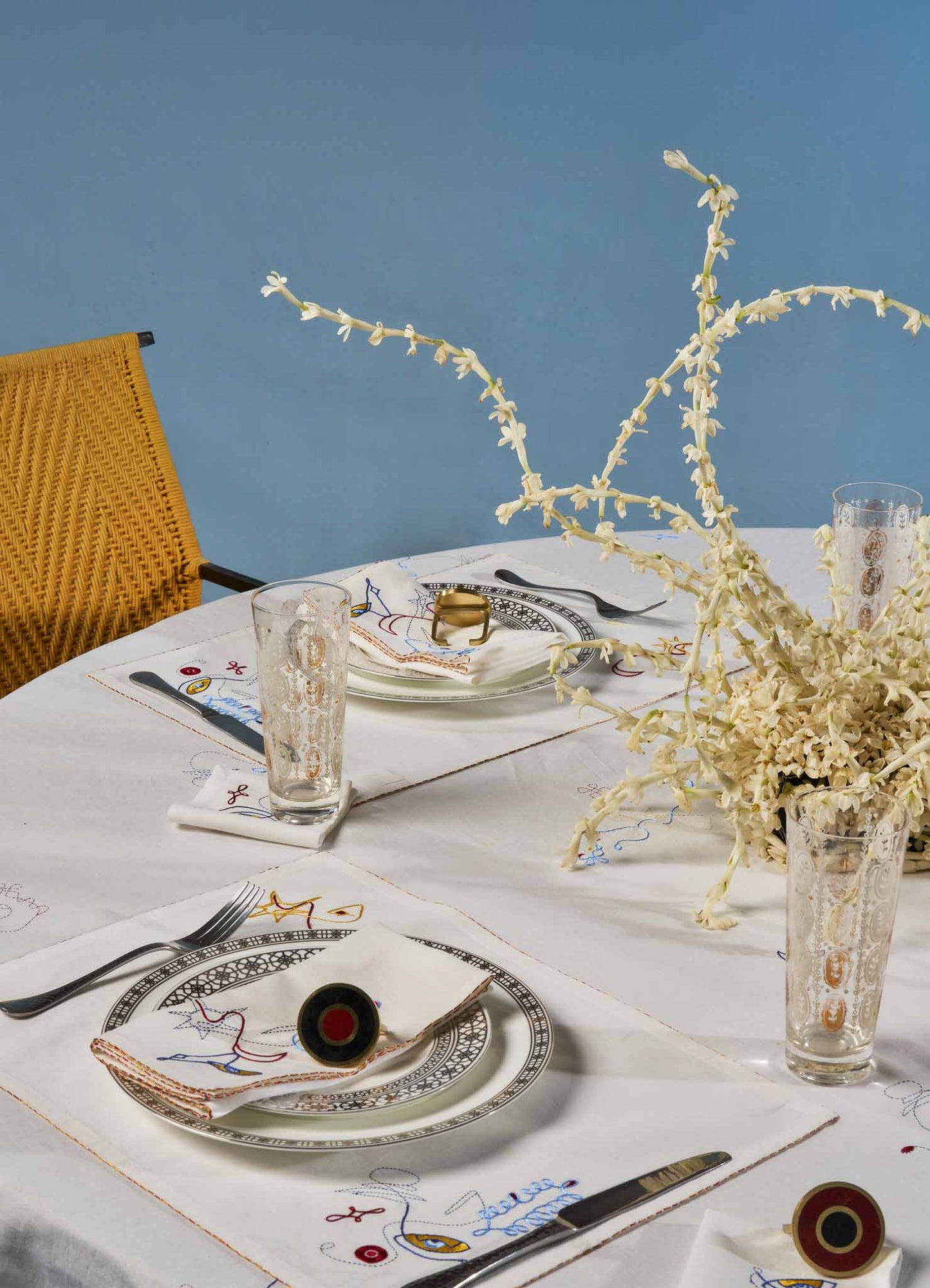 A dining table set with embroidered white tablecloths featuring colorful stitch details, with clear glass tumblers, patterned ceramic plates, and stainless steel flatware. A large floral centerpiece with cream-colored orchids in glass vases adds a natural touch, and a rattan chair with black armrests is positioned at the side.
