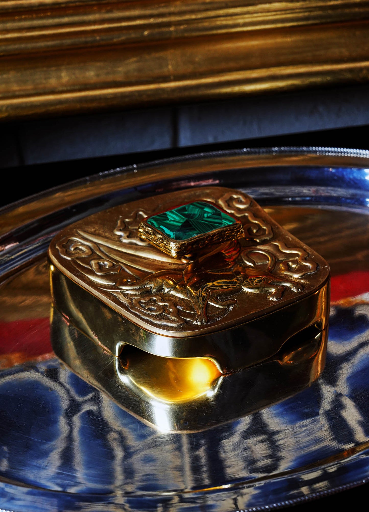 A decorative gold box with intricate embossed patterns and a large green gemstone on top, placed on a reflective silver tray. The box has an antique style with ornate detailing, set against a dark surface background.