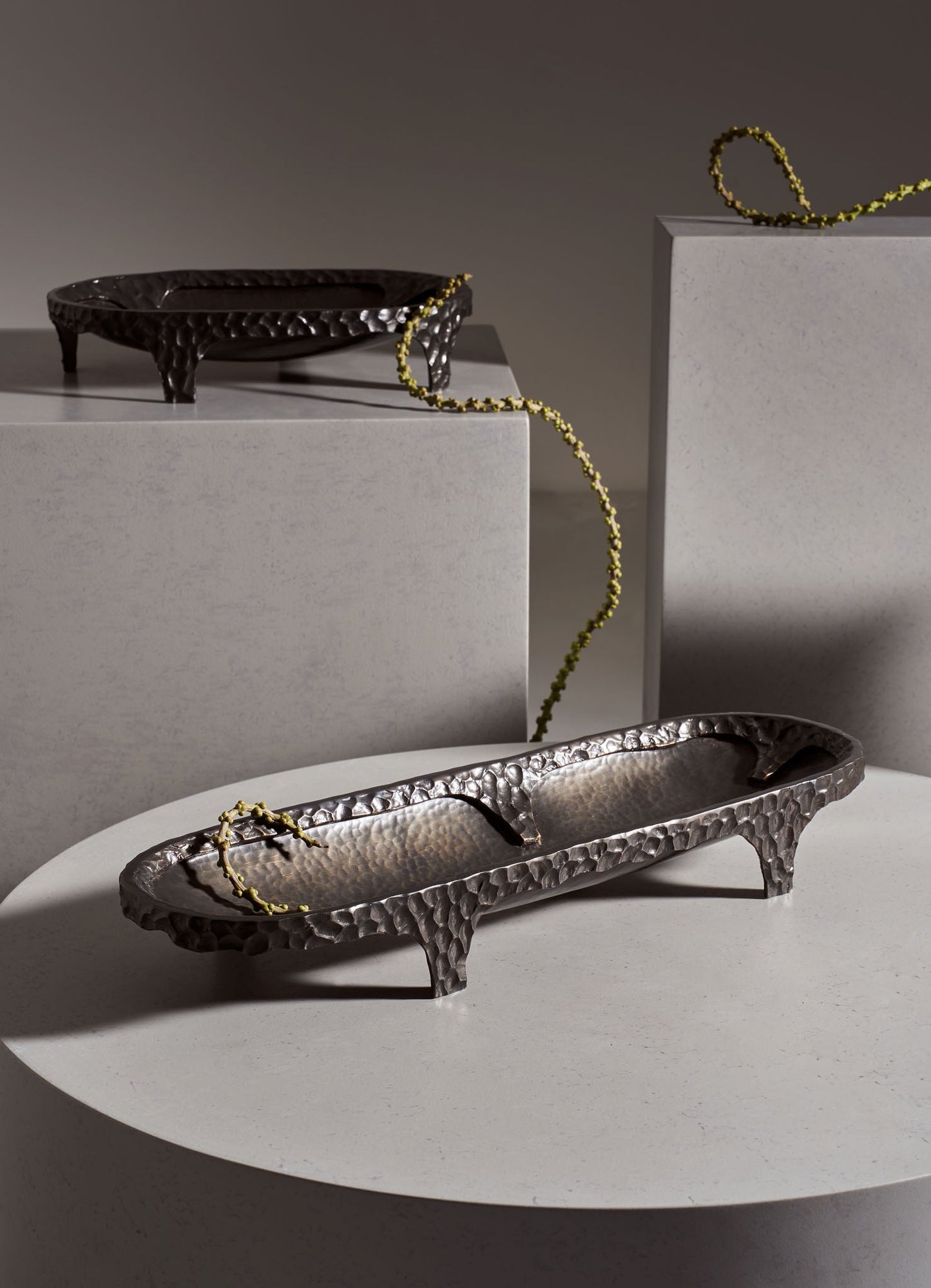 Decorative hammered metal trays with a textured surface and curved edges, finished in a metallic silver tone, displayed on white tabletops with a yellow bead chain draped across them.