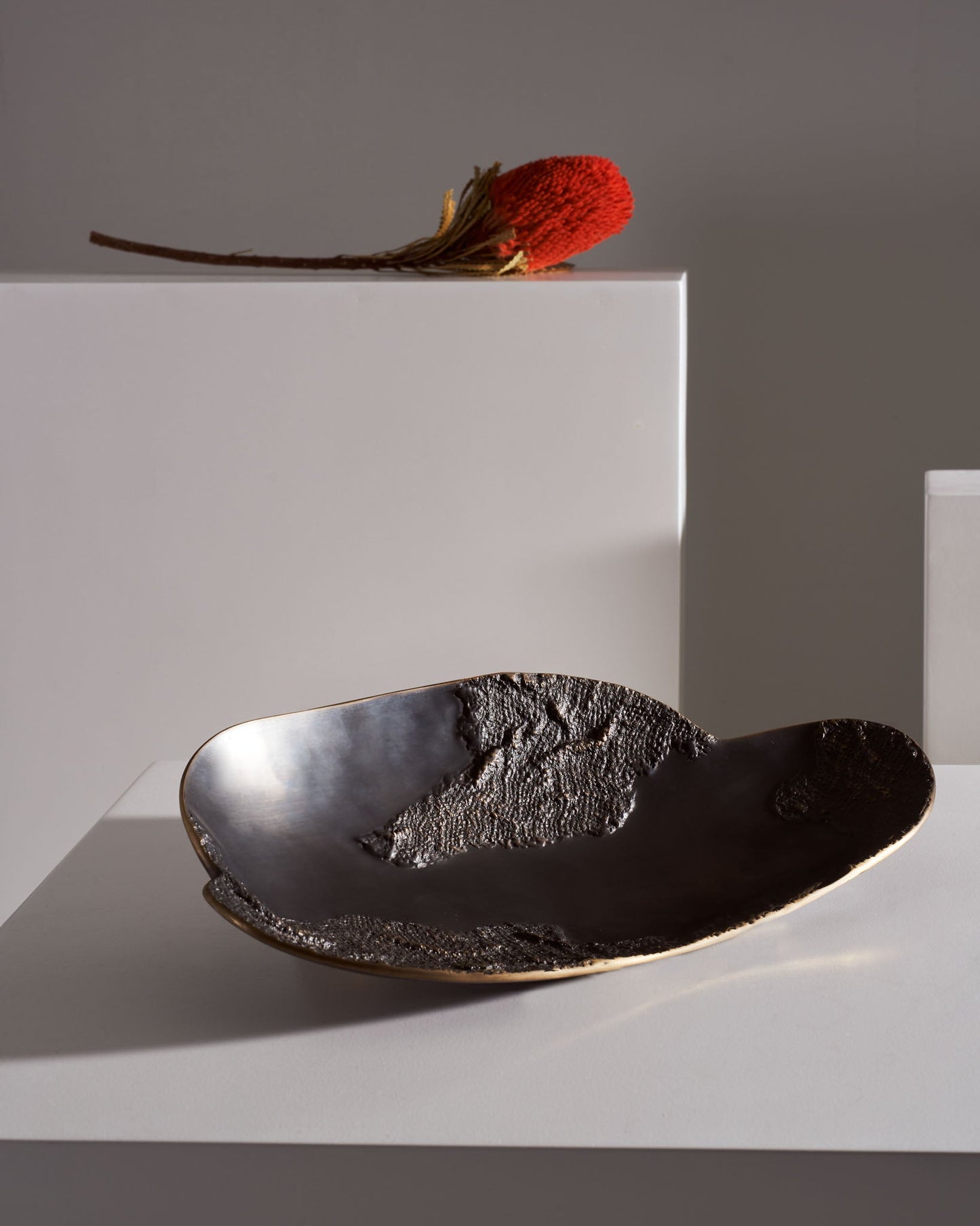 A black metallic bowl with a textured, uneven surface and irregular edges, placed on a white surface with a white geometric block in the background. The overall style appears modern and minimalist.