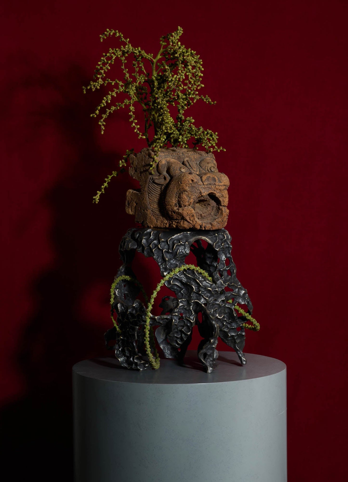 Decorative sculpture with a wooden carved face motif placed on a black, textured metal base, featuring dried green foliage emerging from the top against a dark red background.