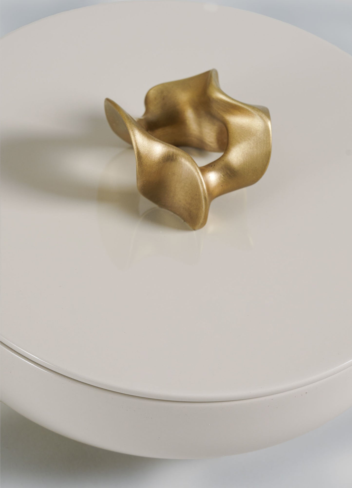 The VIYA Saffron Ecru Trinket Box Large features an abstract, gold-colored sculpture with smooth curves, adding a contemporary touch as it rests atop the glossy, round off-white box.