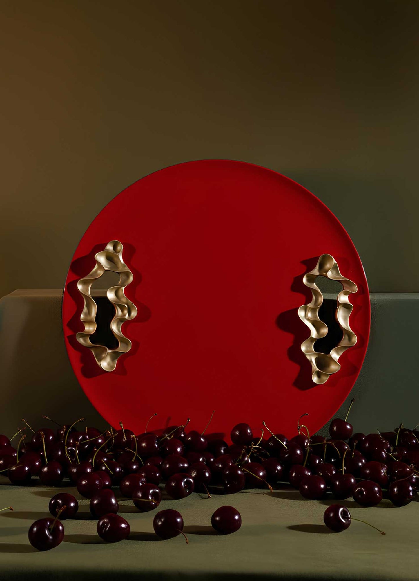 The VIYA Saffron Red Tray Small, featuring two wavy gold handles and a round red design, stands upright behind fresh cherries on a green surface with a muted green background.