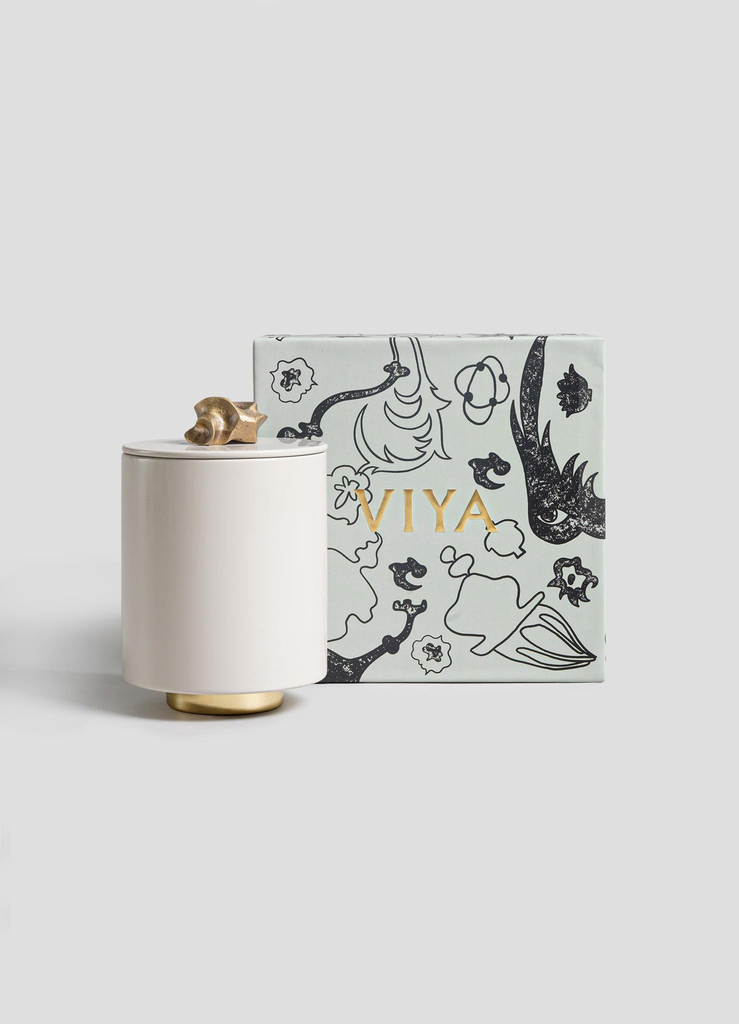 The Saffron Ecru Round Container by VIYA features a white jar with a gold base and snail-shaped lid, beside a light blue box with black abstract designs and VIYA in gold, inspired by bronzed objects and ancient Indian medicine.