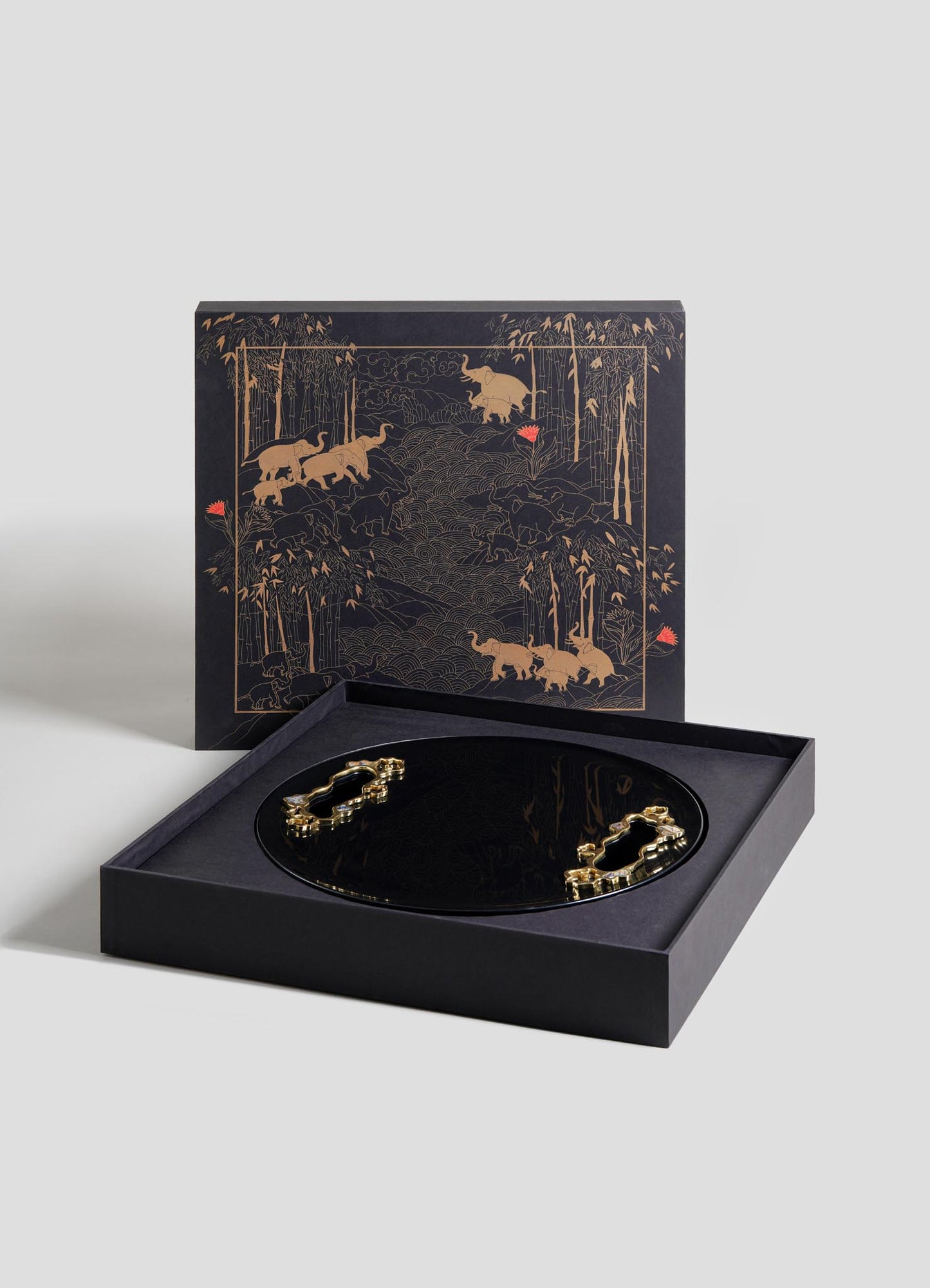 The VIYA Saffron Ecru Tray Large is a round, black tray with gold animal designs, presented in an open box. Its lid displays matching gold animal and forest motifs on a dark background with subtle red accents.