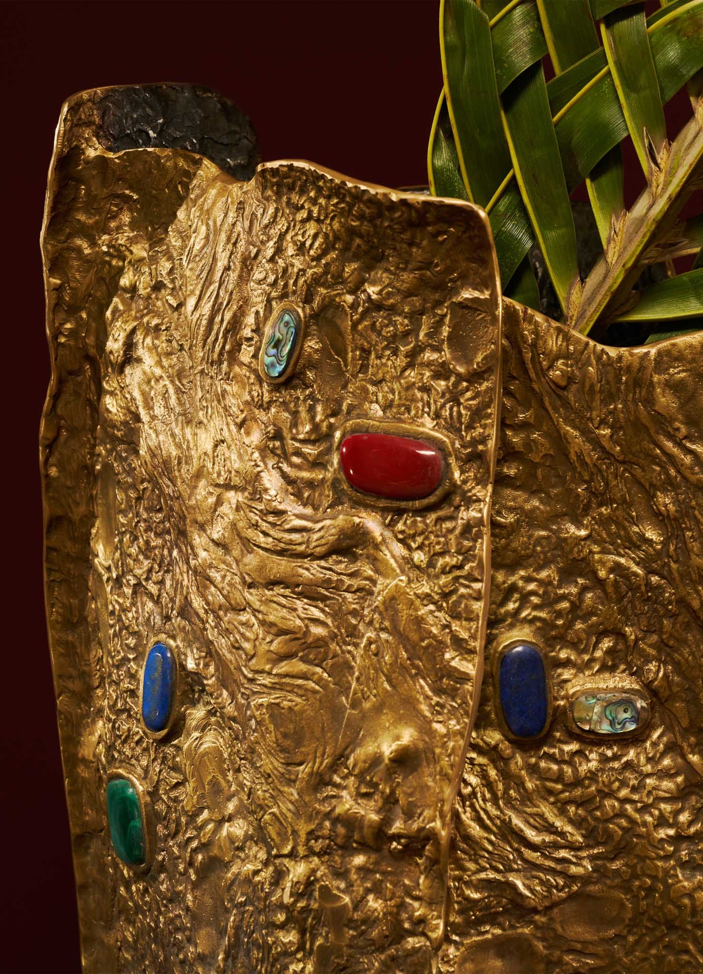 A close-up of a decorative gold-colored ceramic vase with a textured, hammered surface and embedded multicolored gemstone accents in red, blue, green, and an iridescent hue, paired with lush green foliage.