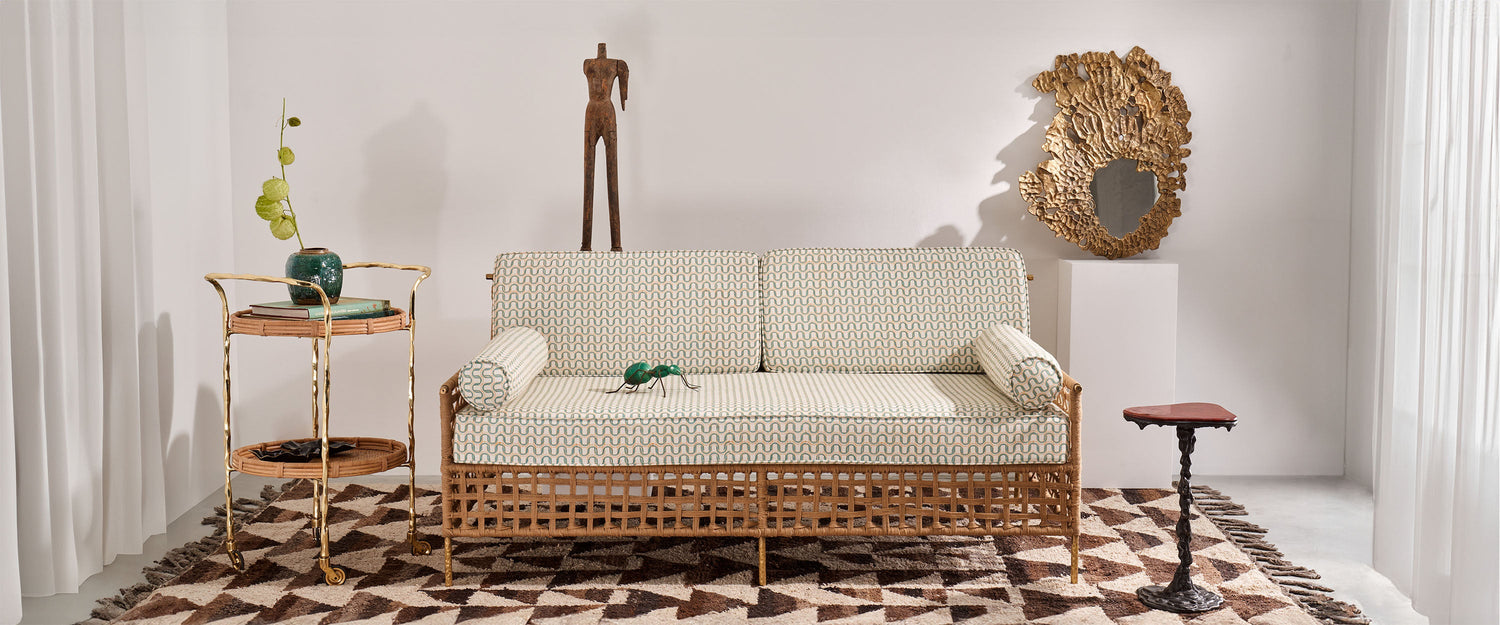A rattan sofa with a woven pattern and light upholstery, accompanied by cylindrical cushions, is set in a contemporary living room with a geometric-patterned wool rug. Decor includes a tall metal statue, a carved wooden side table, a framed mirror with an organic, textured gold frame, and a side table with a metal twisted stem and a wooden top. A green assignment figure is on the sofa, and a vase with green foliage sits on a two-tiered metal and rattan side table.