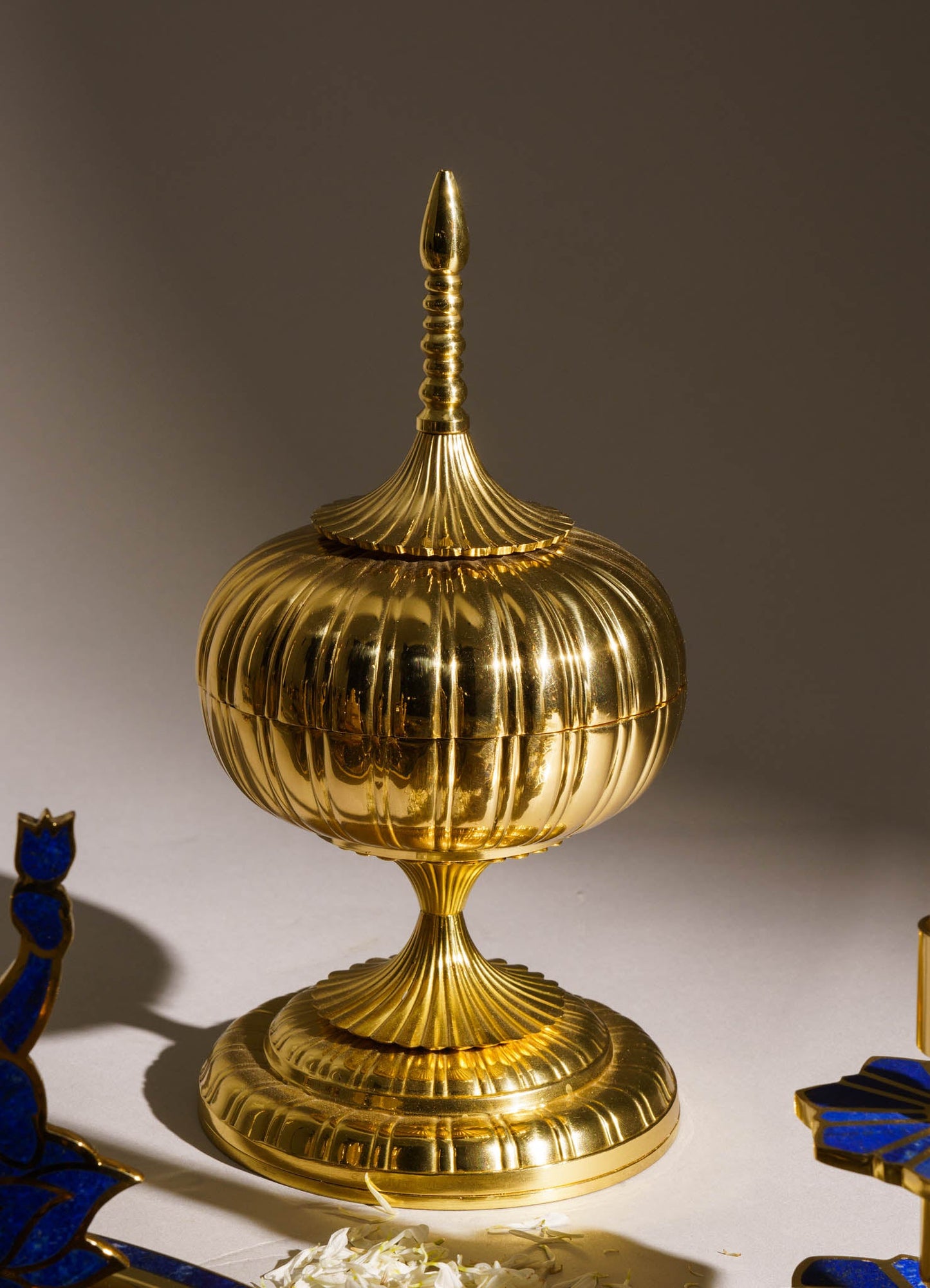A gold-colored ornamental container with a ribbed, rounded body, a tall, pointed finial, and a flared base featuring vertical ridges, exhibiting a decorative, luxury style.