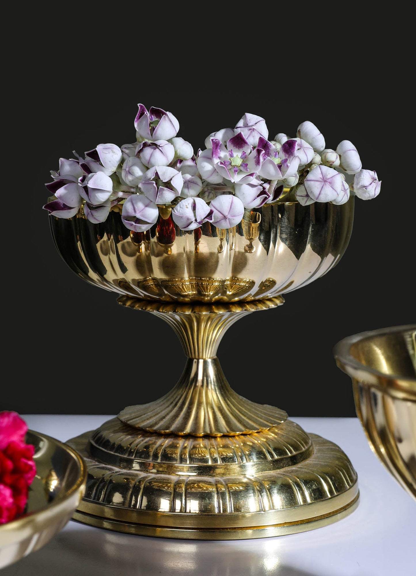A metallic gold pedestal bowl with a fluted base and ribbed design, filled with a cluster of white and purple star-shaped flowers. The bowl has a reflective surface and is displayed on a white surface against a dark background.