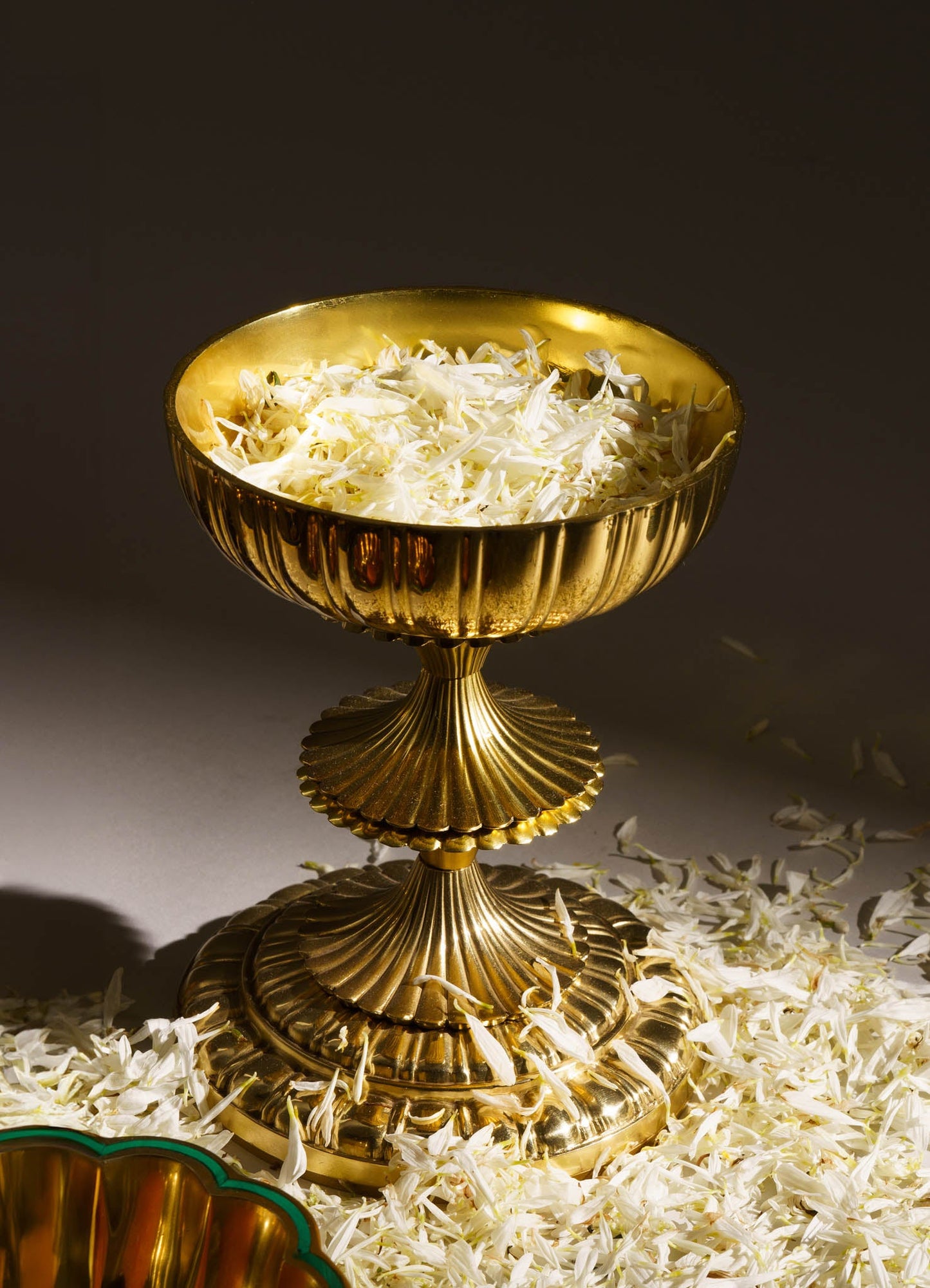 A gold-toned bowl with a ribbed exterior and a fluted pedestal base, filled with white flower petals and placed on a surface scattered with more petals. The bowl has a shiny, reflective finish and a classical style.