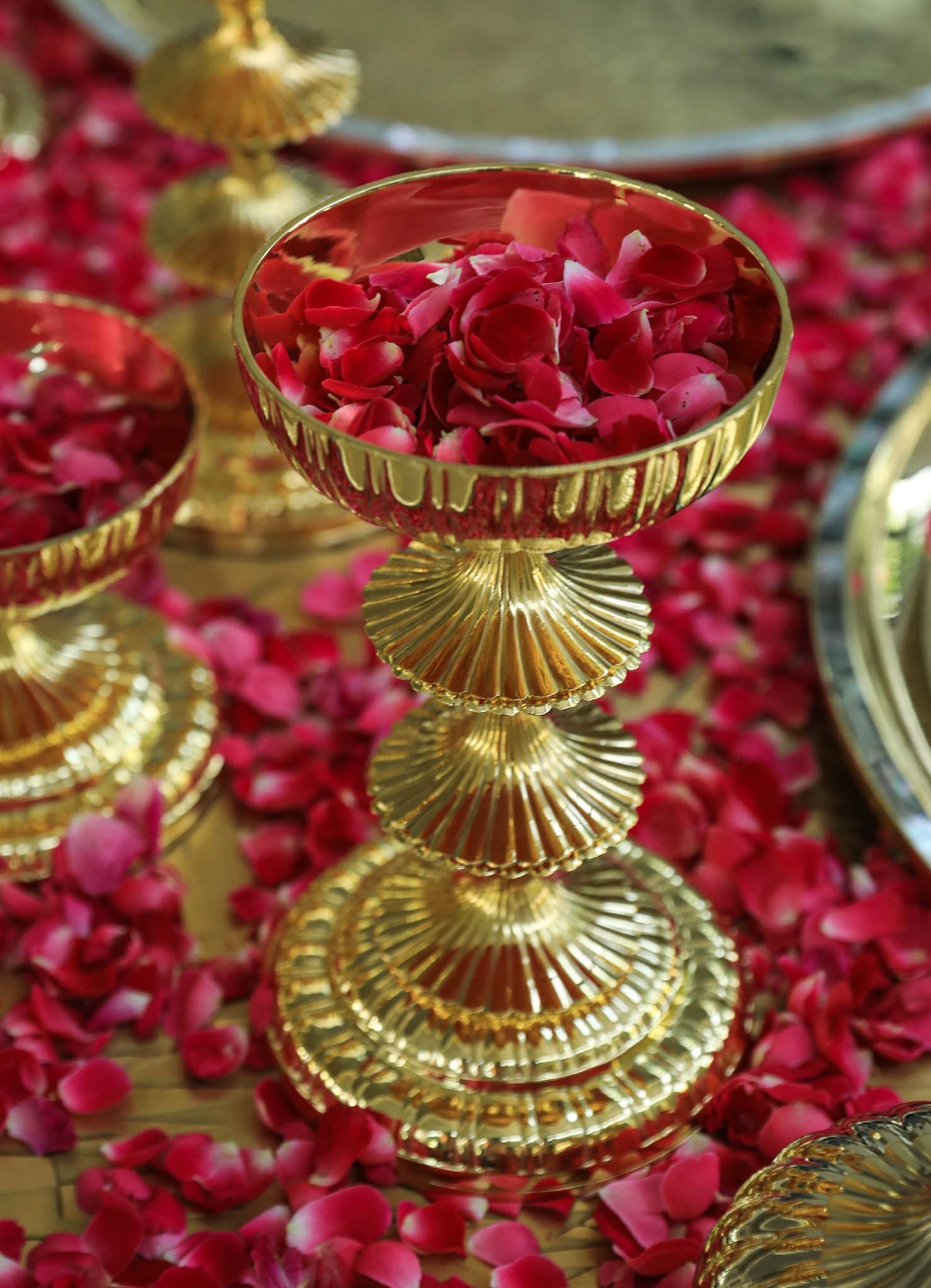 Gold-colored metal pedestal bowls with fluted bases and scalloped edges, filled with pink rose petals, set against a background of matching petals and reflective surfaces.