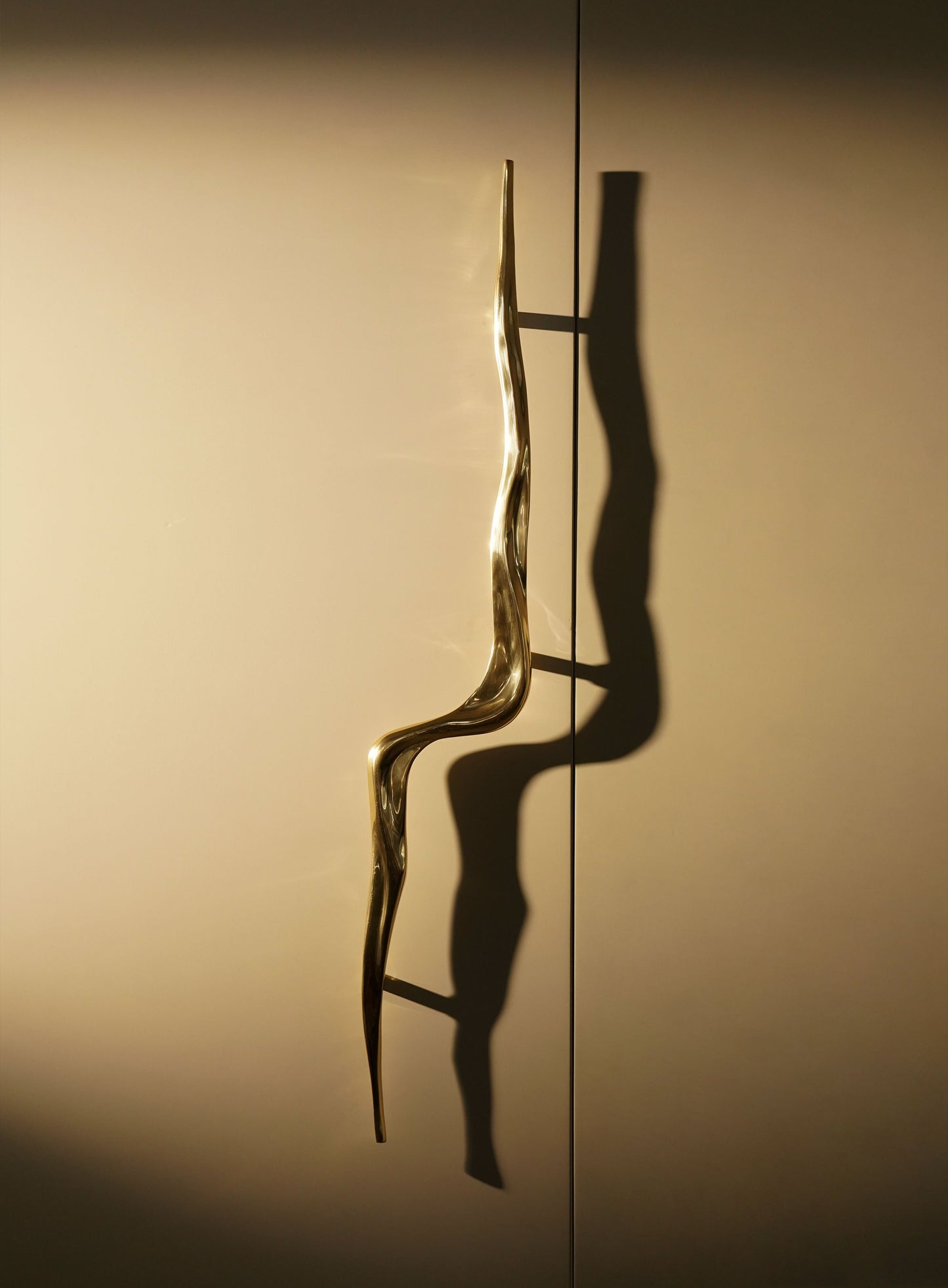A decorative wall-mounted sculpture with an organic, wavy metallic gold finish, casting a shadow on a neutral-colored wall and creating a contemporary, sculptural visual impact.