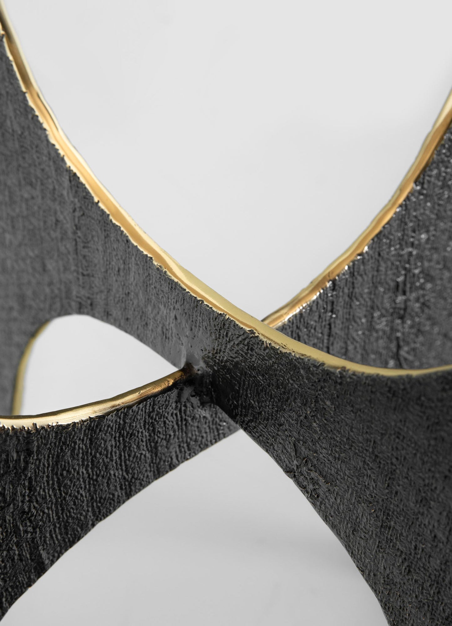Close-up of a black textured metal frame with a gold trim, featuring a modern, sculptural design with intersecting curved elements. The overall style appears contemporary with an emphasis on bold geometry and contrasting finishes.