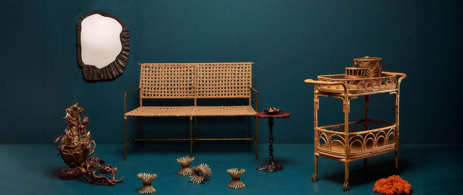 Image features a variety of decorative items, including a rattan shelving unit with curved design details, a woven rattan bench with a metal frame, and several small decorative objects in metal and natural materials. The background is a solid teal wall with a black, irregularly shaped mirror.