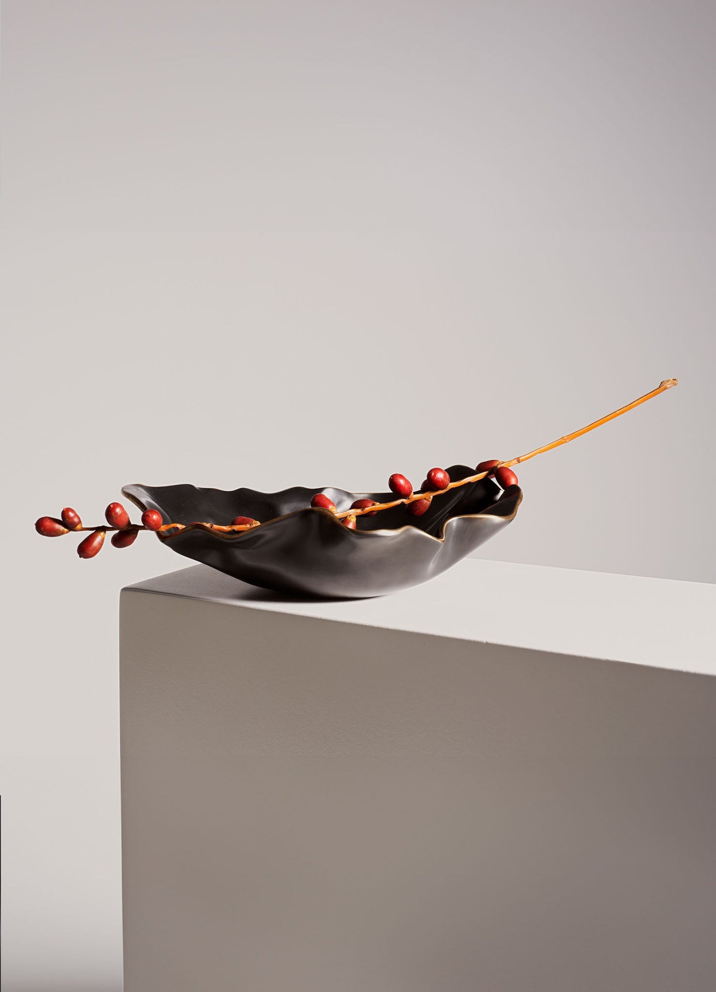 A ceramic bowl with a wavy, irregular edge in matte black, holding a decorative branch with red berries and a thin, elongated stem, set on a plain white surface.