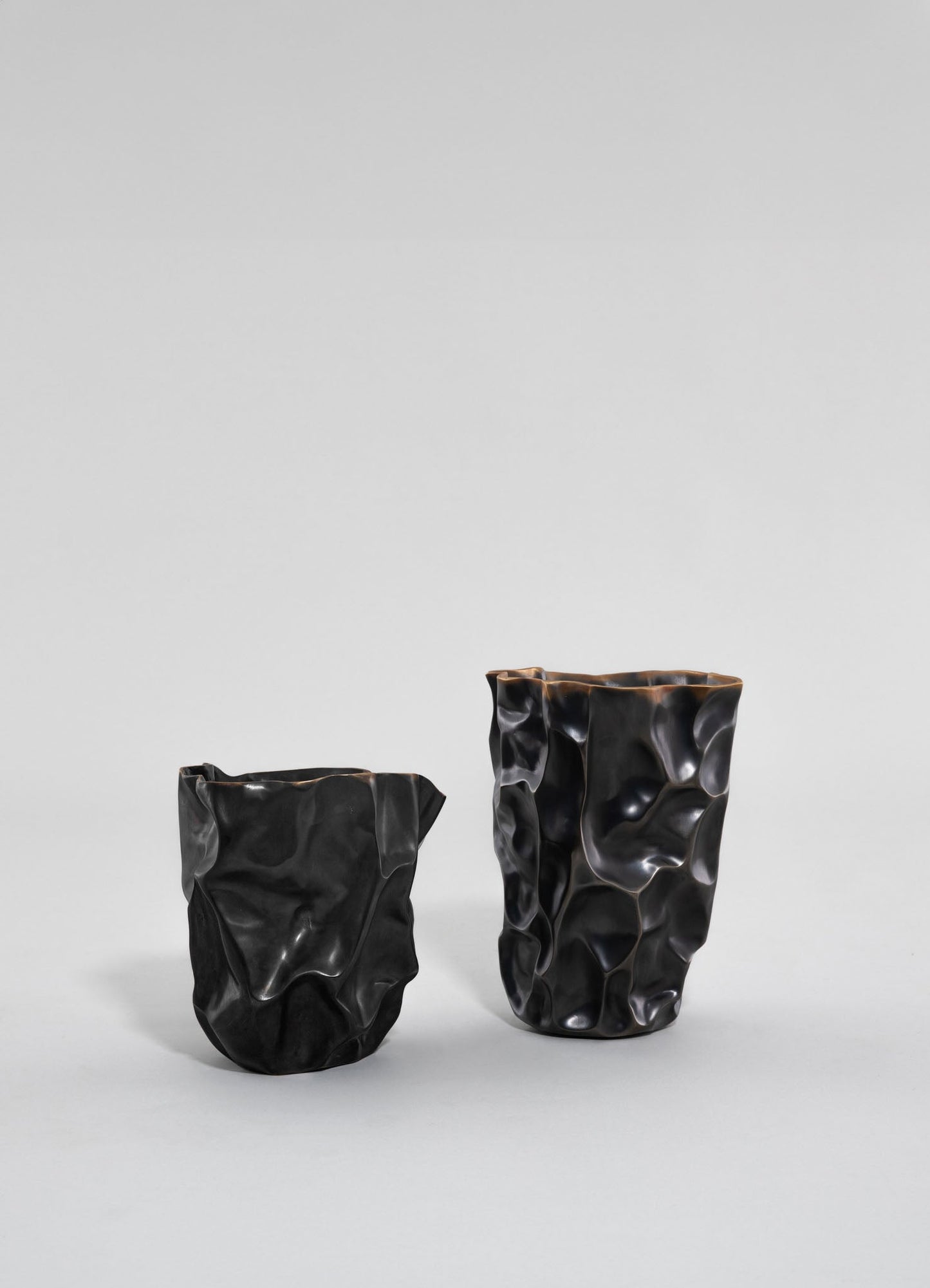 Two sculptural vases with irregular, crinkled surfaces in matte black finish, featuring organic, flowing shapes and contrasting polished edges. The larger vase is positioned to the right of the smaller one, both displayed against a neutral background.