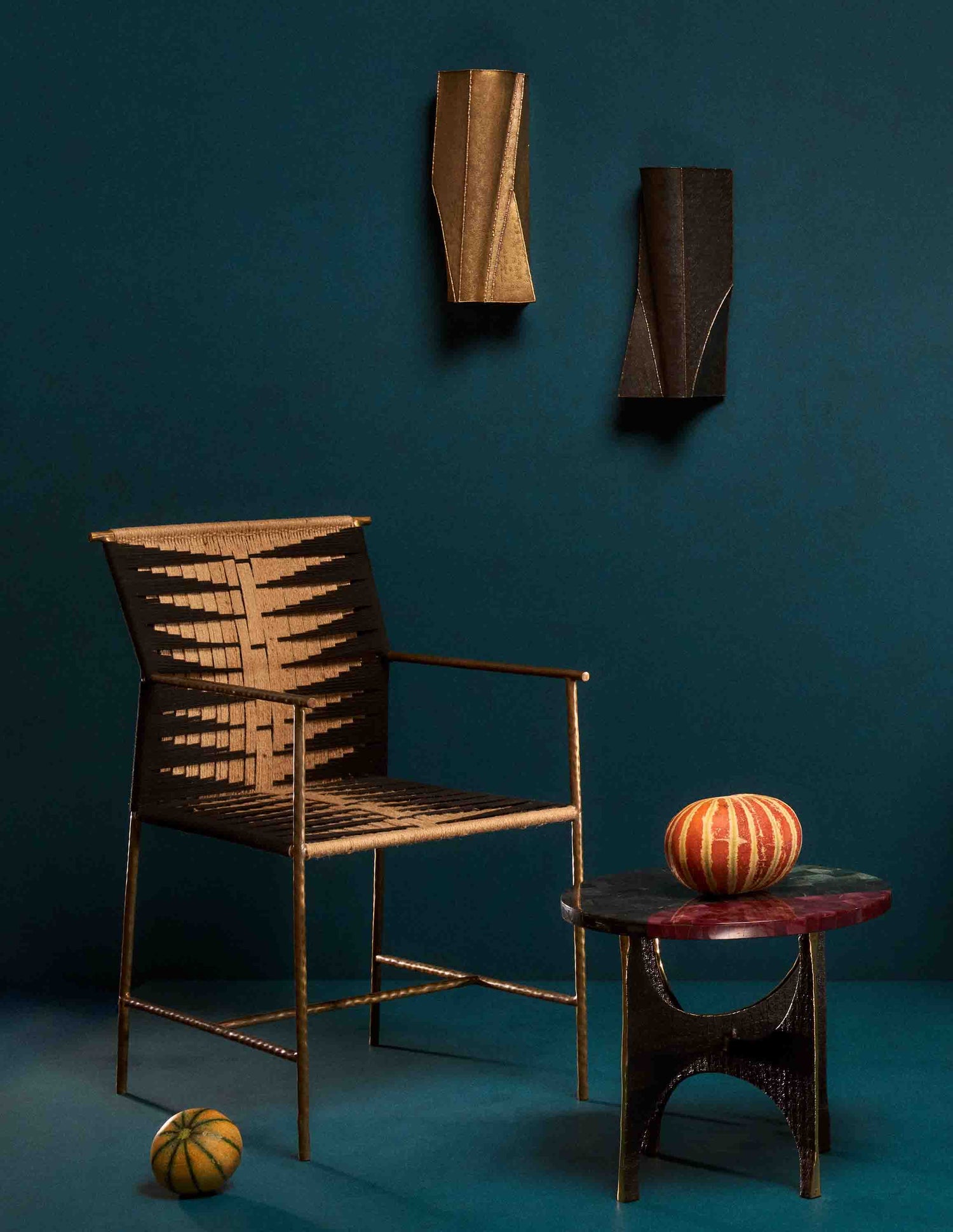 Leather accent chair with a woven seat and metal frame, paired with a small black side table featuring a striped orange and gold decorative object on top, set against a deep teal background with wall-mounted leather and brass wall art.
