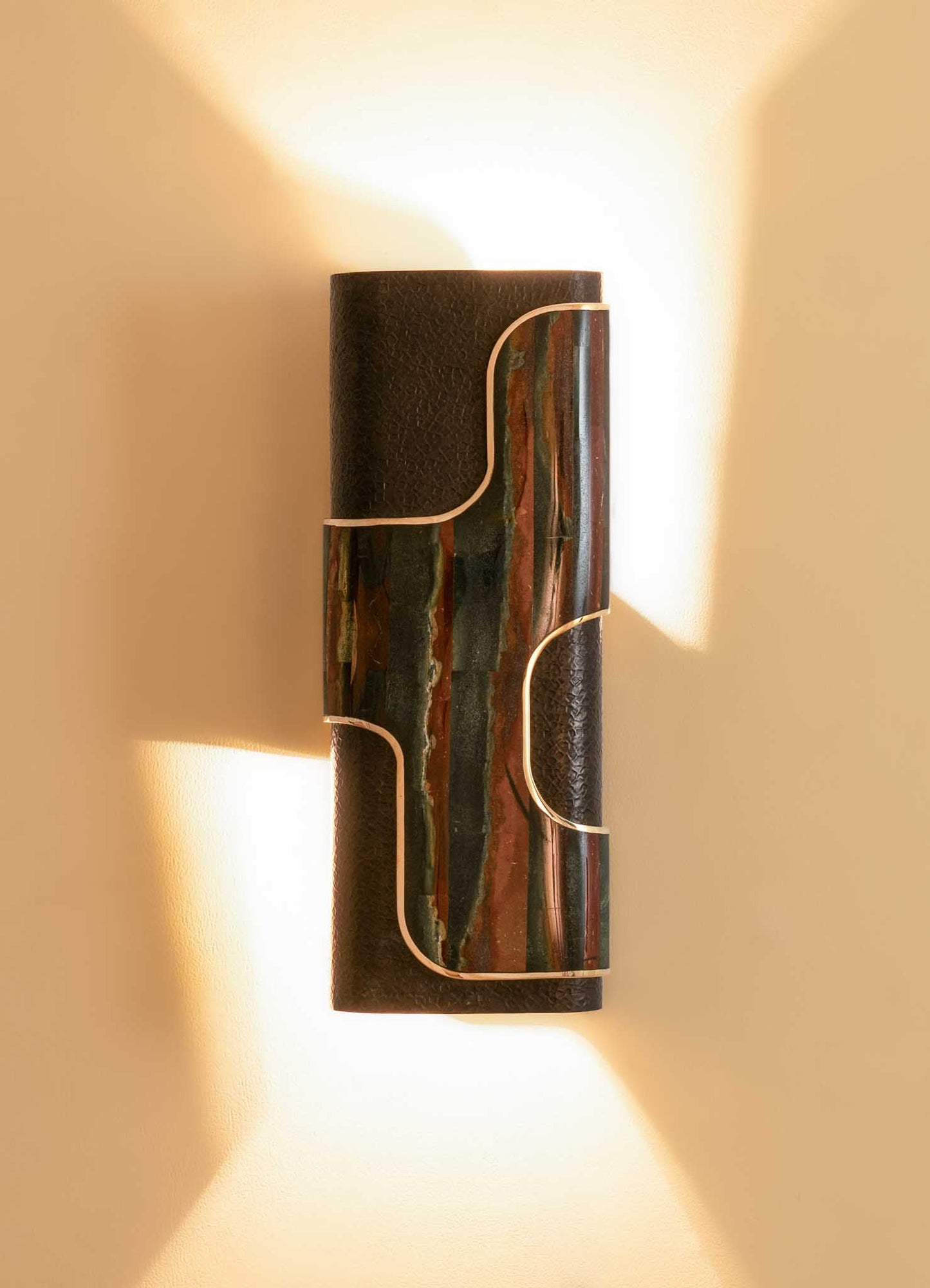 A wall-mounted light fixture with a textured black base and a rectangular centerpiece featuring an abstract pattern of earthy tones, outlined with metallic trim, casting warm light on a neutral wall.