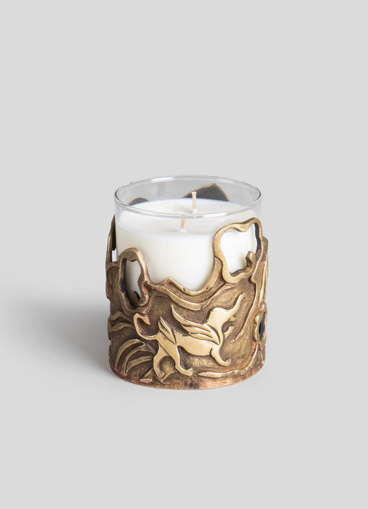 A glass candle holder with a bronze-toned metal sleeve featuring an intricate, swirling dragon motif. The clear glass container holds a white pillar candle and is set against a plain light gray background.