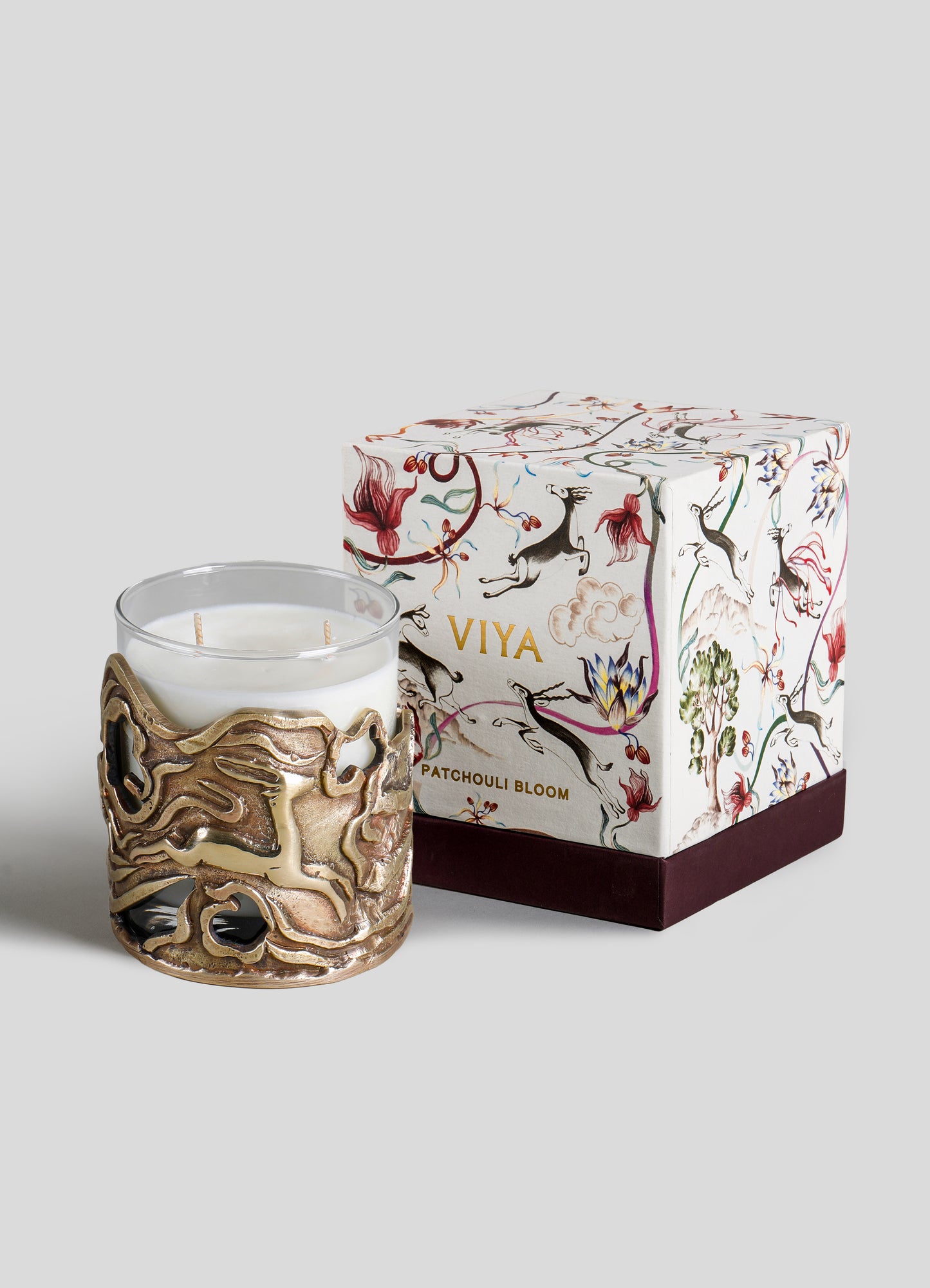 A decorative white candle with two wicks, encased in an ornate gold metal holder with abstract, flowing patterns, placed next to a floral-patterned gift box with illustrations of animals and botanical motifs in red, black, and gold tones.