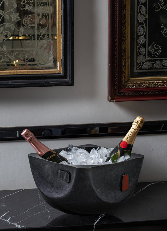 A black hammered metal ice bucket with ergonomic handles holds two bottles of champagne chilling on a dark marble surface, set against a wall with ornate framed artwork and a mirror.