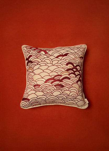 A decorative cushion with a fabric cover featuring a red and beige scalloped pattern, set against a solid red background. The cushion has a square shape with piped edges and a small fabric tag on one corner.