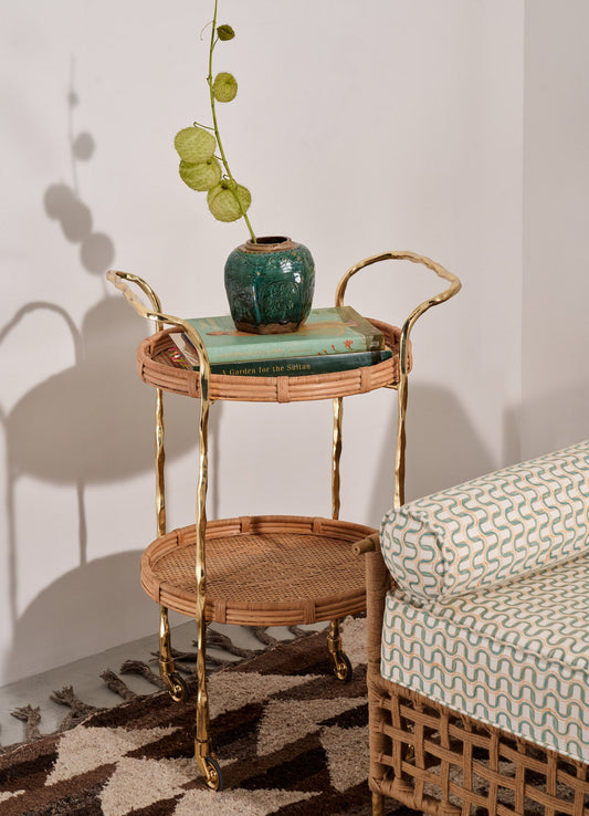 A three-tiered serving cart with a rattan frame and gold-toned metal handles and legs, featuring two round rattan shelves on casters. The scene includes a green ceramic vase with textured surface and spherical botanical accents, and a stack of books, set against a plain wall with a patterned rug on the floor.