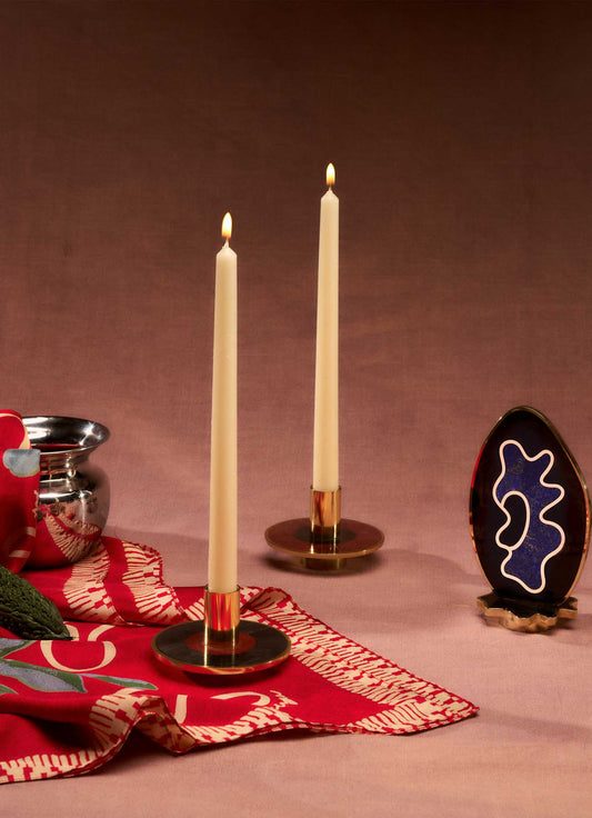 Brass candle holders with a circular base and polished gold finish, holding tall white taper candles with lit flames, set on a pink surface with a patterned red and white textile.