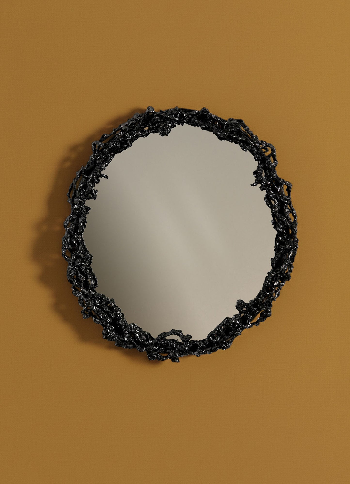 The Andaman Mirror by VIYA, featuring an ornate, irregular black frame inspired by organic forms, hangs on a mustard brown wall and casts a subtle shadow to the left.