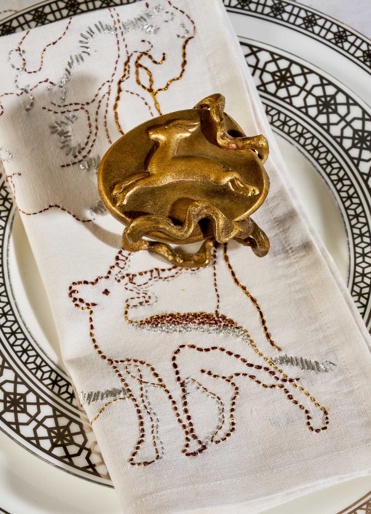 A white napkin with embroidered animals rests on a patterned plate, held by VIYA’s Blackbuck Napkin Ring—gold-toned and shaped like a blackbuck in motion, echoing art from 17th-century Indian manuscripts. Set of 2.