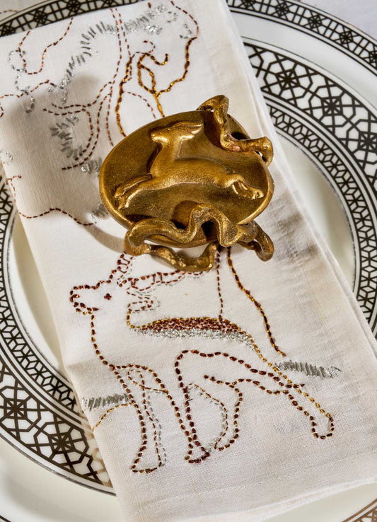 A white napkin with embroidered animals rests on a patterned plate, held by VIYA’s Blackbuck Napkin Ring—gold-toned and shaped like a blackbuck in motion, echoing art from 17th-century Indian manuscripts. Set of 2.
