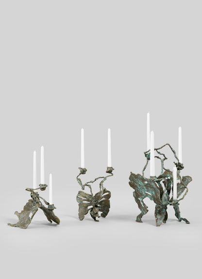The VIYA Butterfly Candle Stand Large features an abstract, sculptural bronze design with organic, twisted forms, each holding two or three tall white taper candles. Displayed against a plain light gray background.