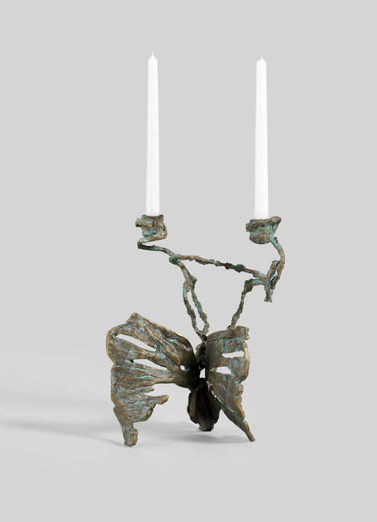 The Butterfly Candle Stand Medium by VIYA is a sculptural metal candlestick holder with an abstract, organic form and a bronze-green patina. It holds two tall white taper candles and features a textured surface set against a plain gray background.
