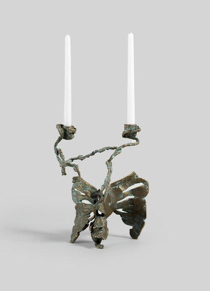 The VIYA Butterfly Candle Stand Medium features a sculptural metal design resembling an abstract butterfly, holding two white taper candles with twisted arms extending from the wings, set against a light gray background.