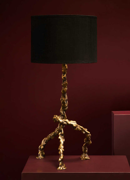 A table lamp with a black fabric drum shade and a sculptural base in a textured metallic gold finish, set against a deep red background.