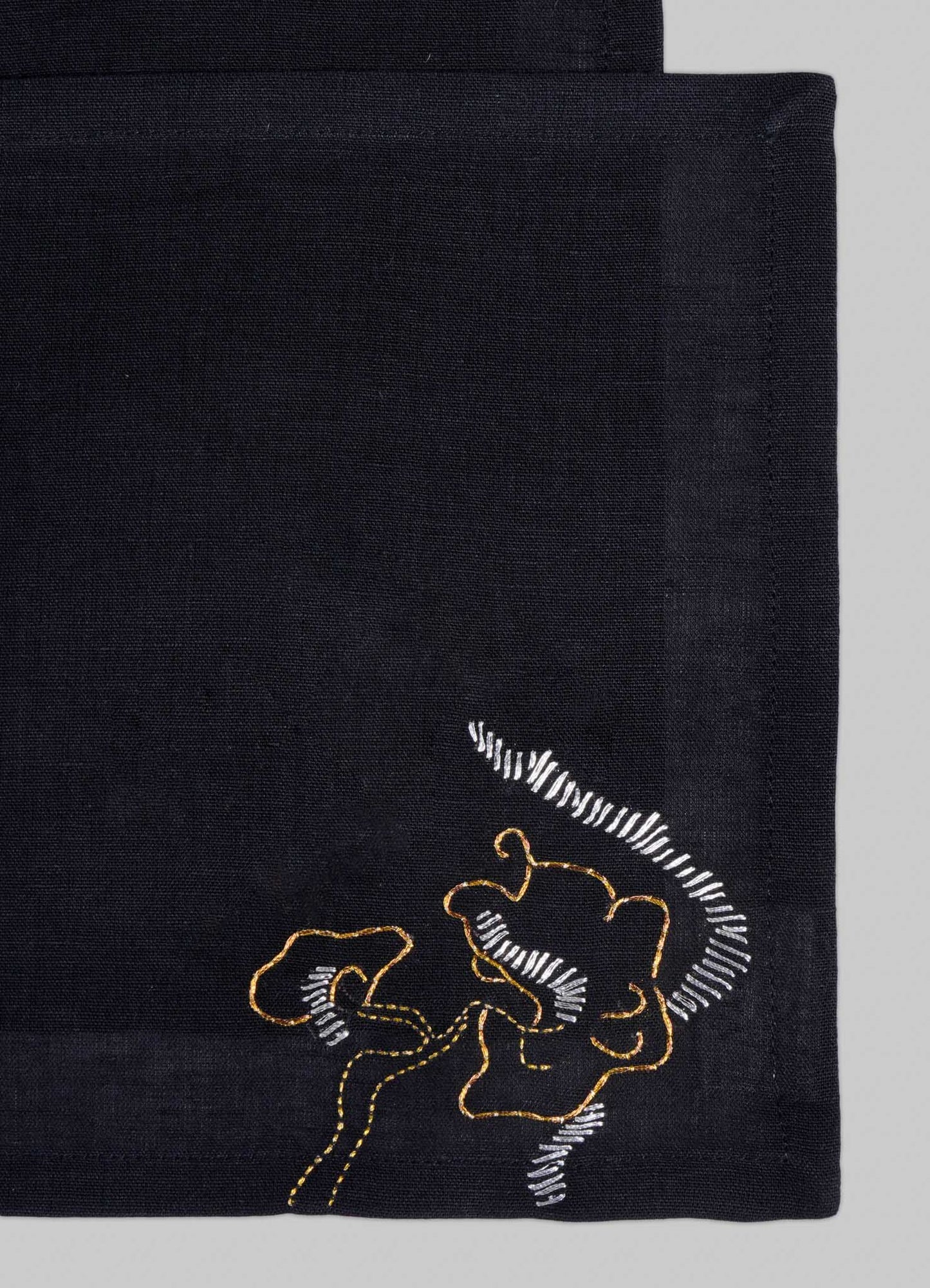 The Viya Blackbuck Dreams Hand Embroidered Black & Gold Cocktail Napkins (Set of 4) feature abstract face motifs in yellow and white thread on black fabric, inspired by Indian manuscripts.