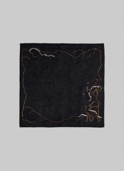 The Viya Blackbuck Dreams Hand Embroidered Black & Gold Napkin features a black square cloth with gold and white abstract embroidery inspired by Indian manuscripts, including a stylized human figure in the lower right corner.