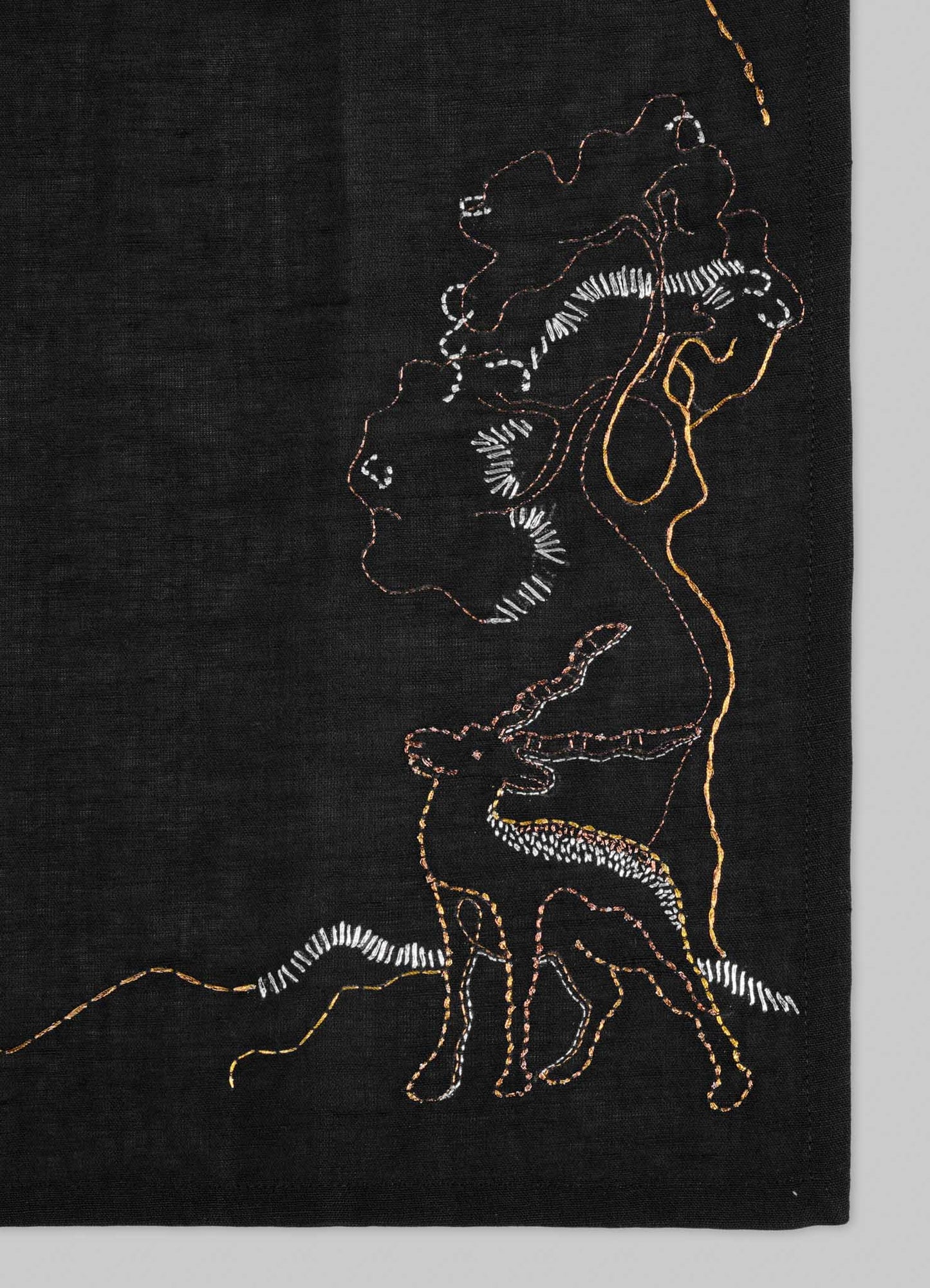 The Viya Blackbuck Dreams Hand Embroidered Black & Gold Napkin features colorful outlines of a woman with a vessel, a deer, and a tree on black fabric, stitched in white, orange, and yellow for an Indian manuscript-inspired look.