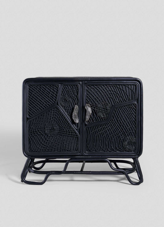 The VIYA Chakra Cabinet features two front doors with geometric cane weaving, curved legs, and two clear handles, all in black rattan, set against a plain light gray background.