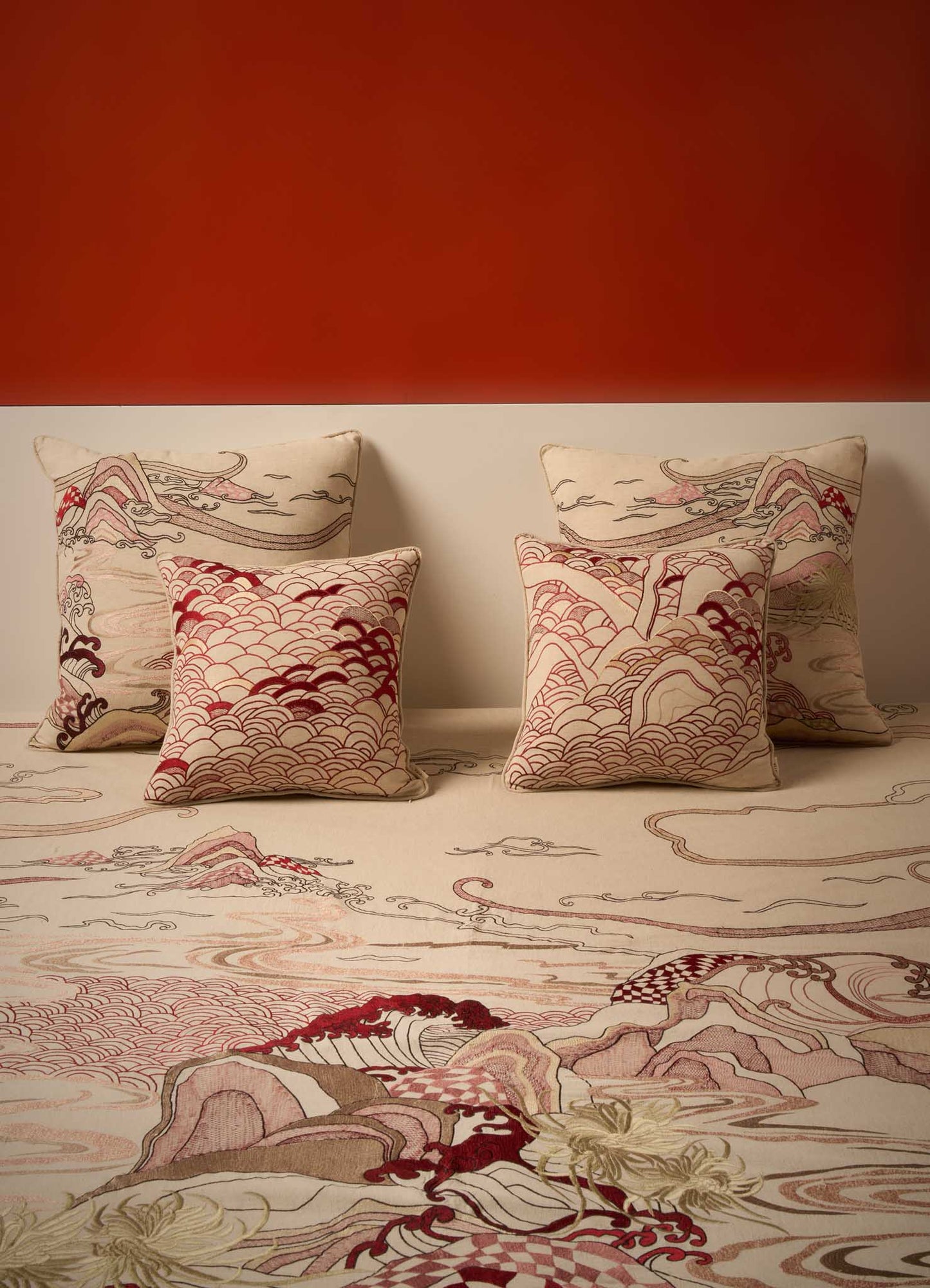 Four Viya Dreamscape Red Embroidered Cushion Covers (24"x24") featuring intricate wave and mountain motifs in beige, red, brown, and cream are arranged against a muted wall with a matching carpet and a bold burnt orange background.