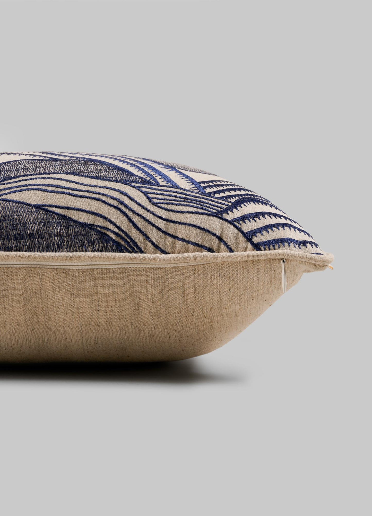 A close-up side view of the Viya Dreamscape Blue Embroidered Mounds Cushion Cover (12"x18") shows its beige fabric with blue geometric motifs and a visible zipper along the edge, set against a plain gray background.