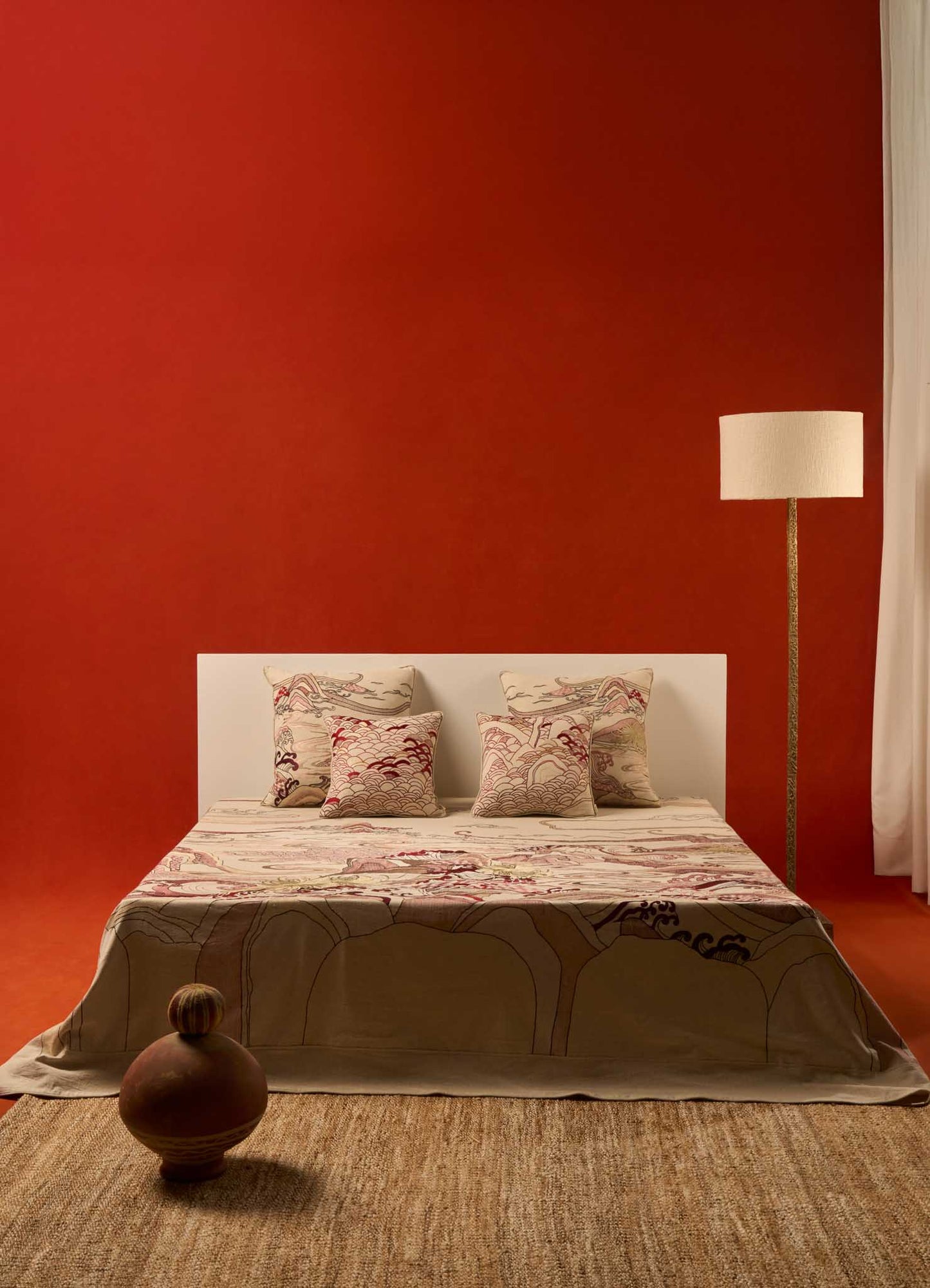 A minimalist bedroom features the Viya Dreamscape Ecru Embroidered Bedcover on a white bed set against a bold red wall, earth-toned pillows, symbolic motif textiles, tall floor lamp, clay vase, woven rug, and white curtains to the right.