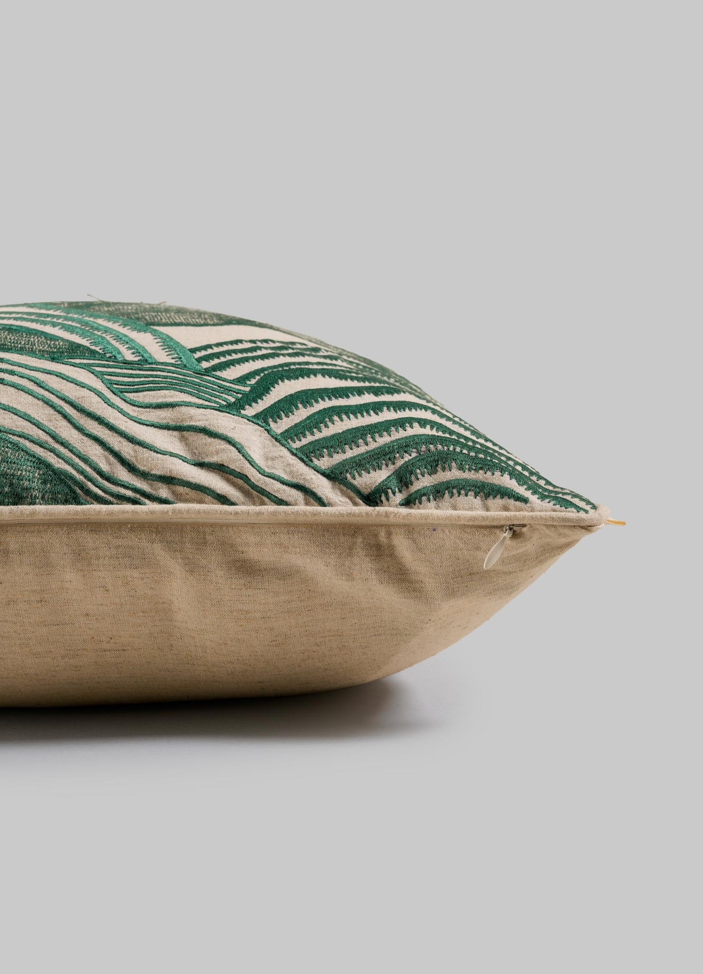 A close-up side view of Viya’s Dreamscape Green Embroidered Mounds Cushion Cover (18"x18") features Rajput manuscript-inspired motifs on beige fabric with a visible edge zipper, set against a plain gray background.